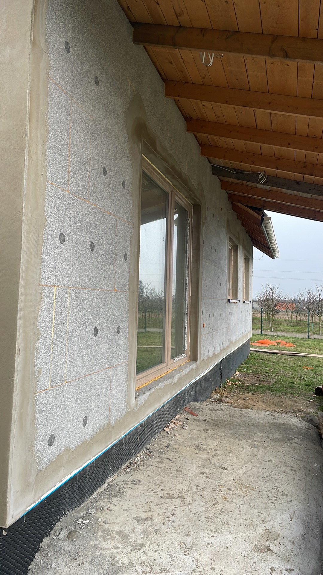 Elewacja budynku w trakcie ocieplania, widoczne płyty styropianowe z charakterystycznymi okrągłymi wgłębieniami, częściowo pokryte warstwą kleju, okna z drewnianymi ramami, drewniane zadaszenie...