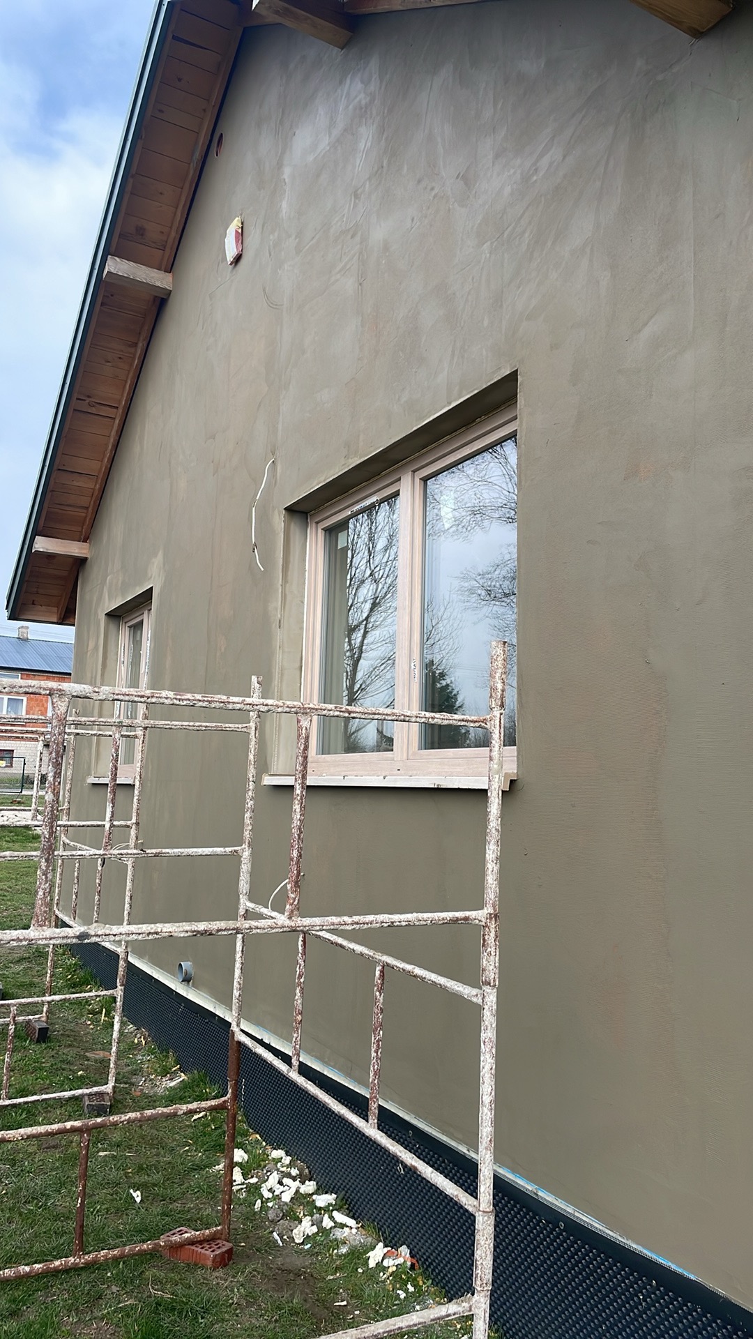 Świeżo otynkowana ściana zewnętrzna domu jednorodzinnego w kolorze beżowym, widoczne rusztowanie, okno z białymi ramami i fragment dachu z drewnianą podbitką.