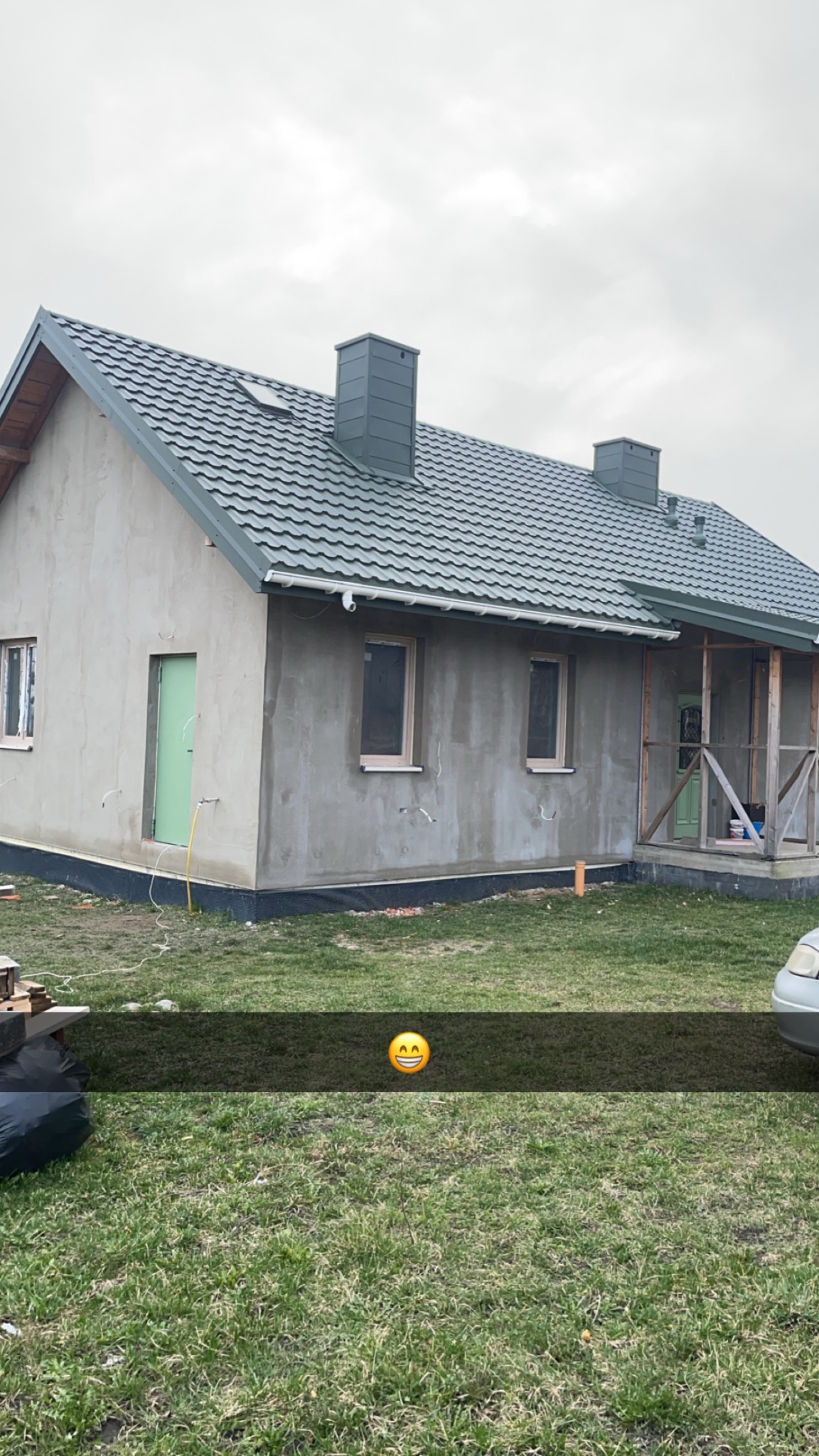 Dom jednorodzinny w trakcie prac elewacyjnych: surowa szara elewacja, zielone drzwi wejściowe, szara dachówka, widoczne przewody i rurki instalacyjne przy ścianach, trawnik przed budynkiem...