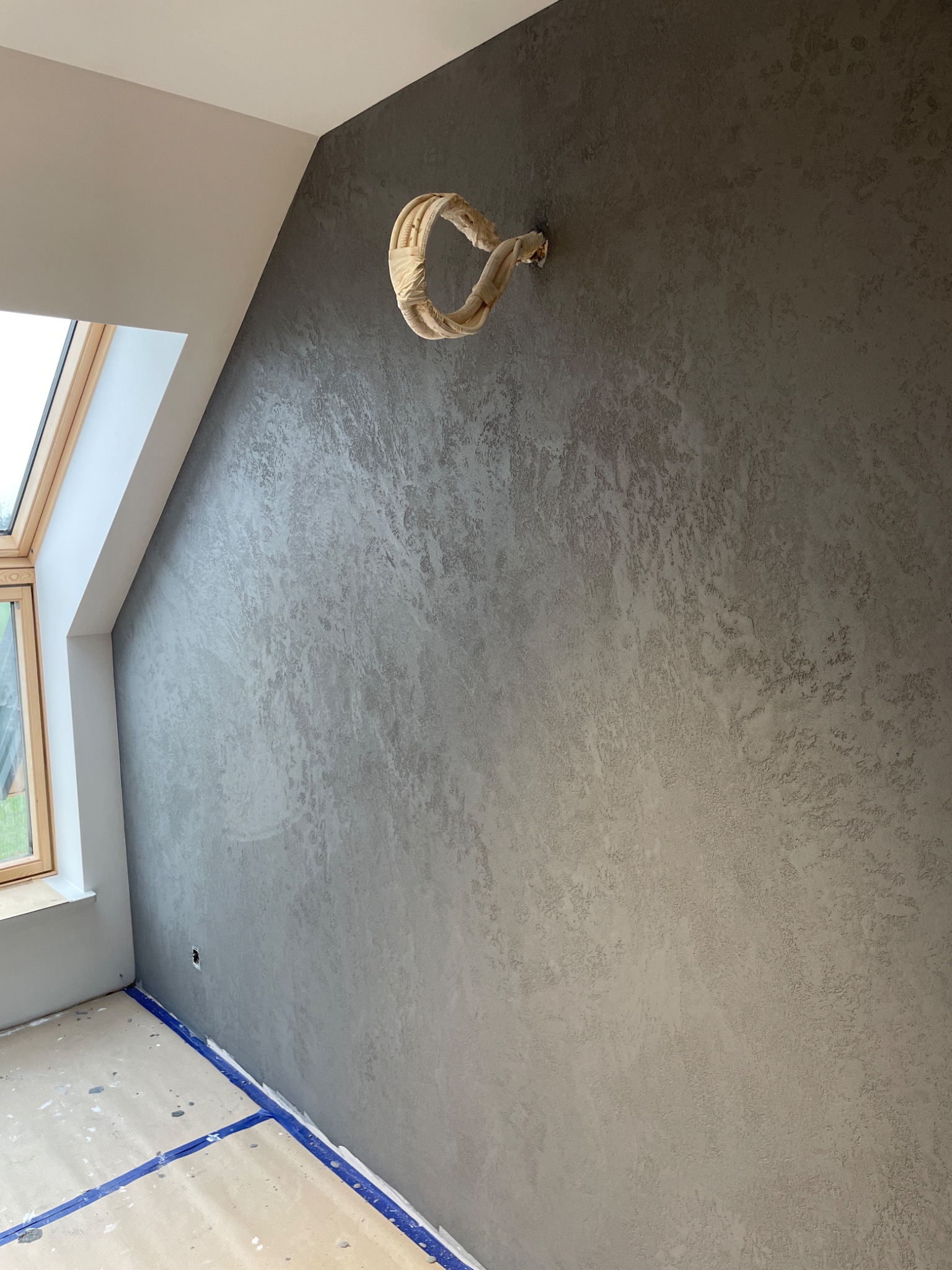 Ściana w pokoju na poddaszu z dekoracyjną szarą strukturą, zabezpieczone rury grzewcze, okno dachowe i taśma malarska na podłodze.
