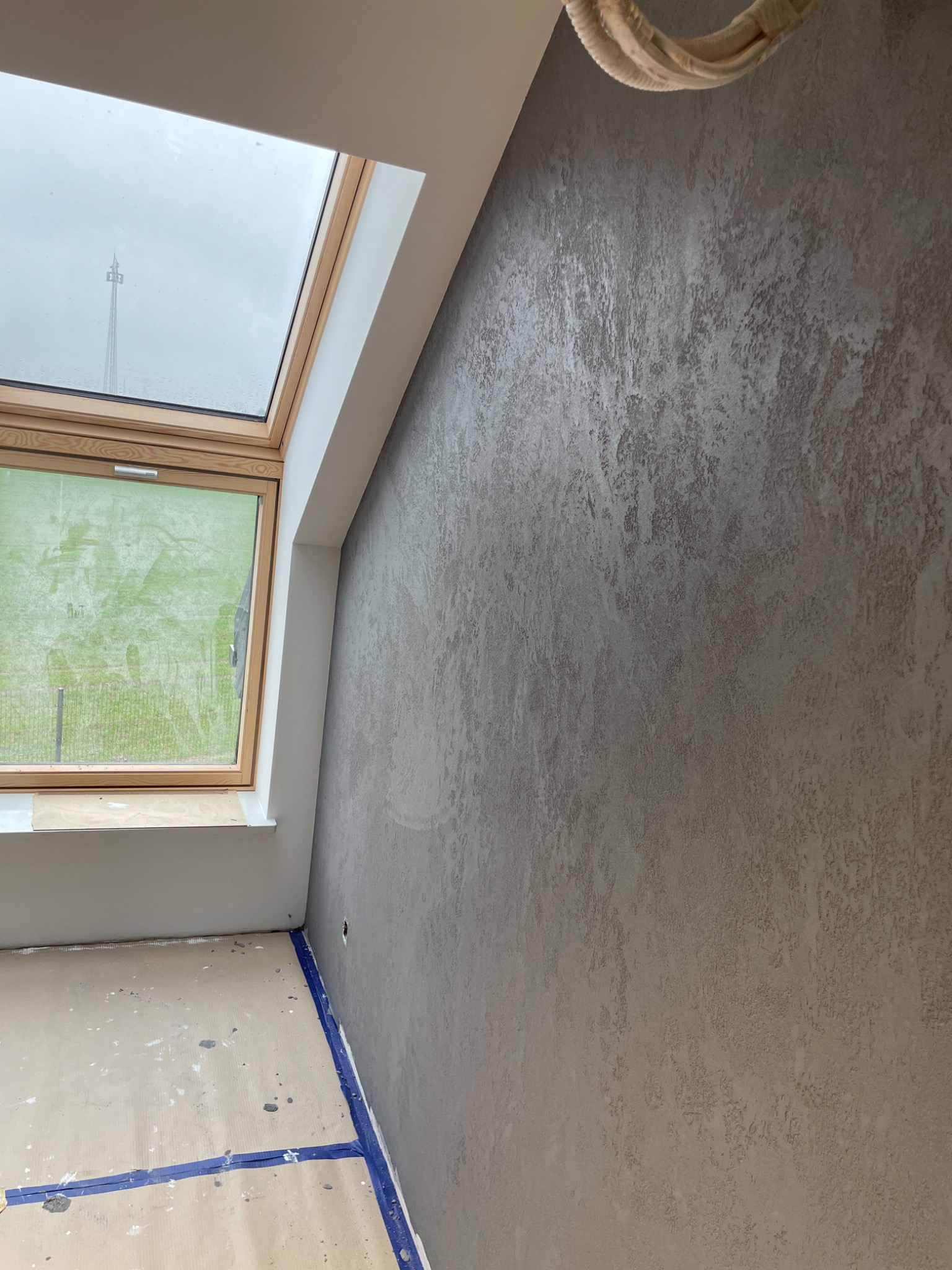 Ściana z dekoracyjną, szarą fakturą w pokoju z oknem dachowym i zabezpieczoną podłogą, widoczne przewody pod sufitem.