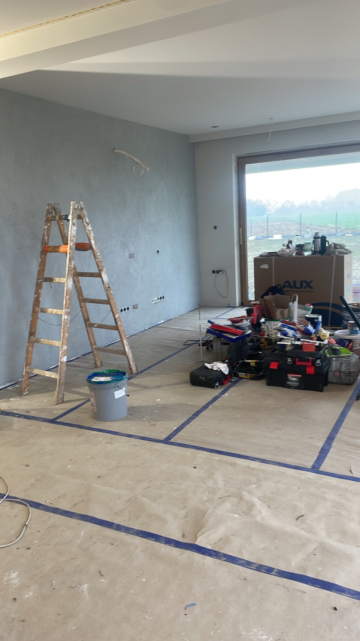 Pokój w trakcie prac wykończeniowych: ściana pokryta szarą strukturą, drewniana drabina oparta o ścianę, podłoga zabezpieczona folią i taśmą, narzędzia i materiały budowlane zgromadzone w rogu...