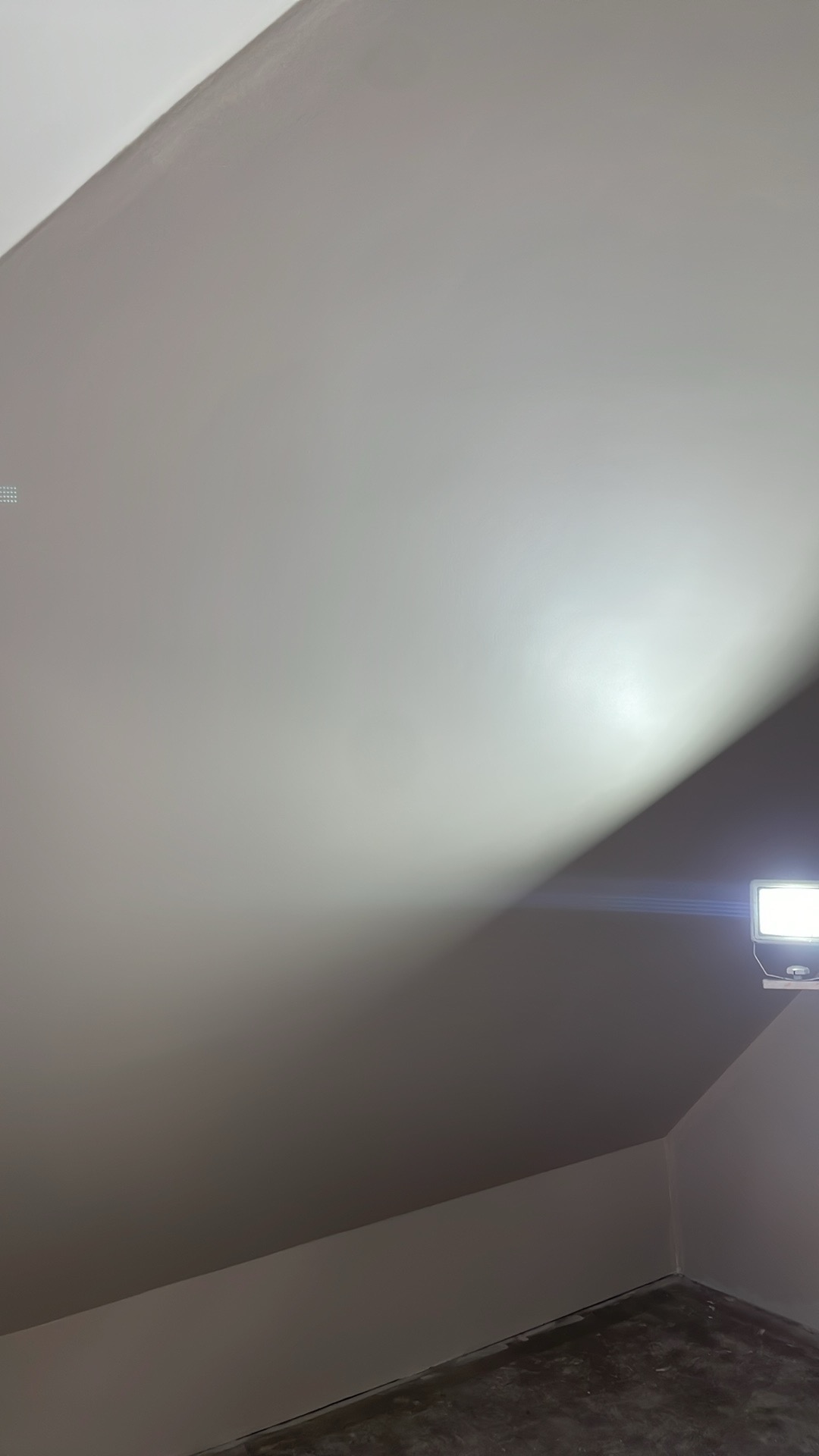 Poddasze ze skośnym sufitem, oświetlone lampą halogenową, ściany świeżo pomalowane na jasny odcień szarości, widoczny fragment surowej podłogi.