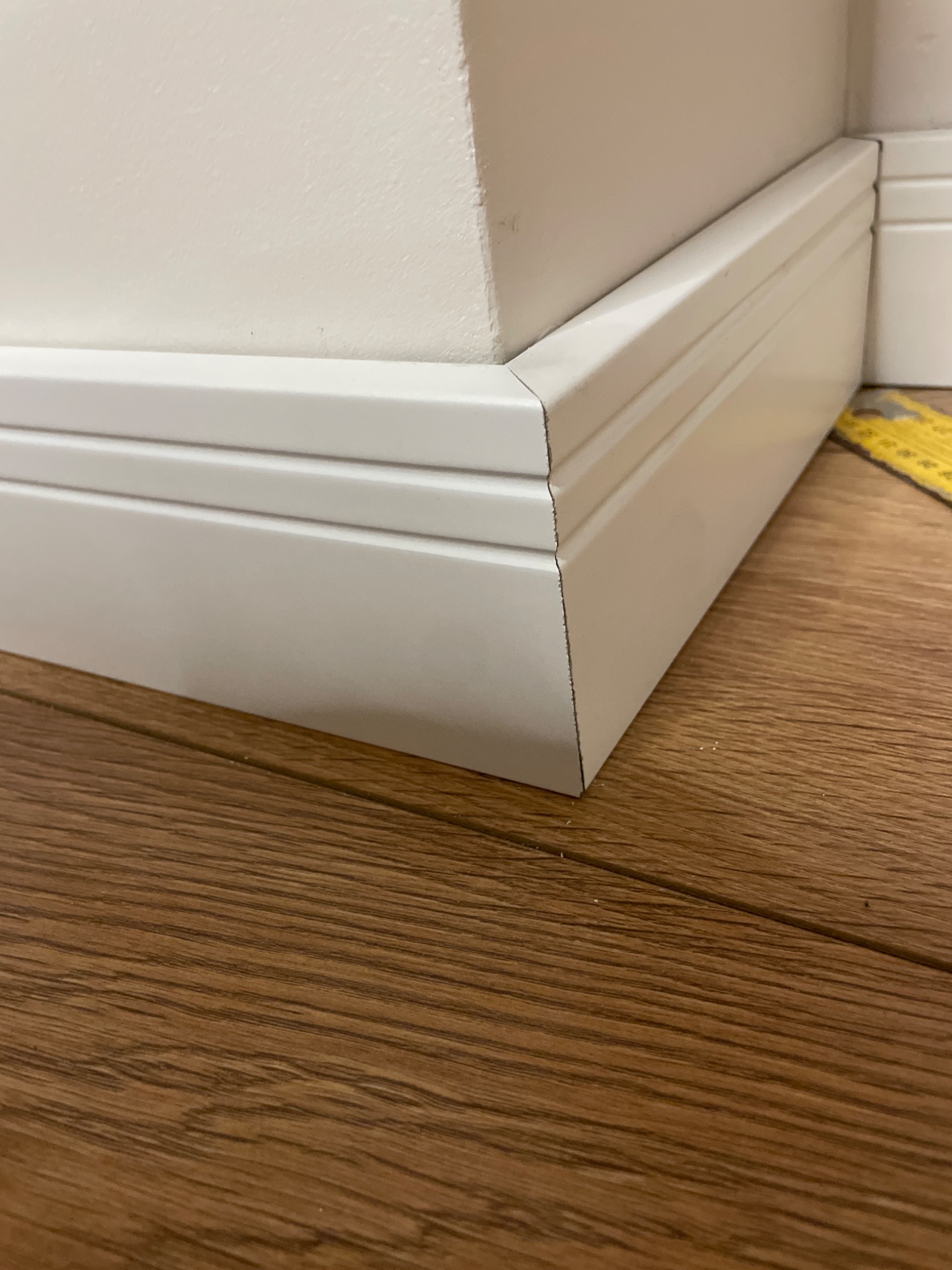 Detal wykończenia: biała listwa przypodłogowa, trójwarstwowa, zamontowana przy ścianie na drewnianej podłodze, z widoczną żółtą miarką w tle.