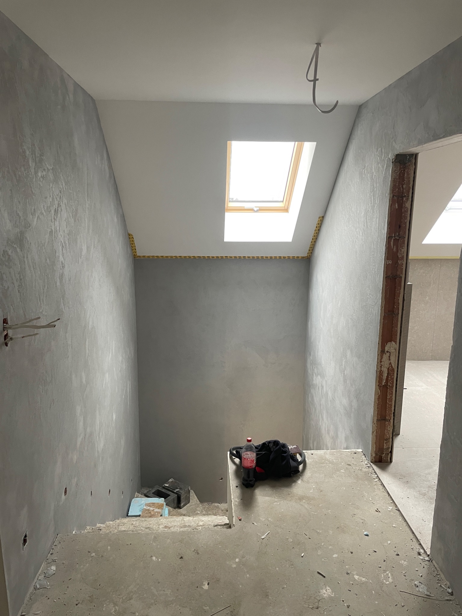 Klatka schodowa w trakcie prac wykończeniowych: ściany pokryte szarą, dekoracyjną strukturą, zabezpieczone żółtą taśmą przy suficie z oknem dachowym, widoczne przewody elektryczne wystające ze...