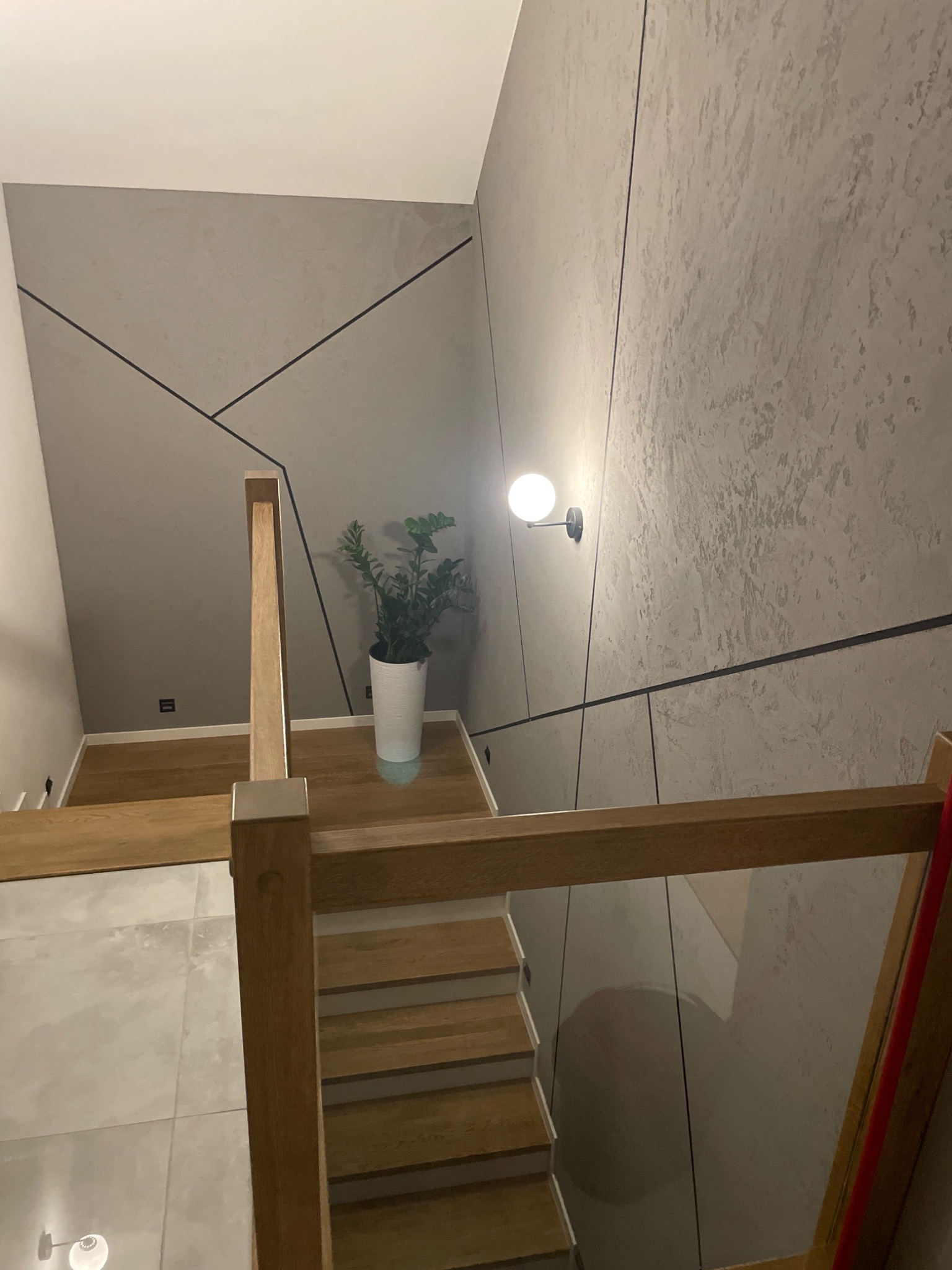 Nowoczesne schody drewniane z jasnymi stopniami, szara ściana z geometrycznym wzorem i lampą kulą, doniczka z rośliną na podeście.