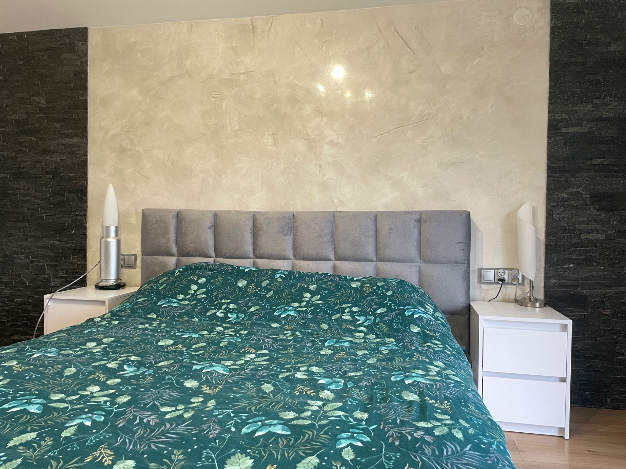 Sypialnia z łóżkiem z zieloną pościelą w roślinny wzór, szarym zagłówkiem i ścianą wykończoną w dwóch stylach: jasny tynk dekoracyjny imitujący marmur i ciemna ściana z kamienia dekoracyjnego.