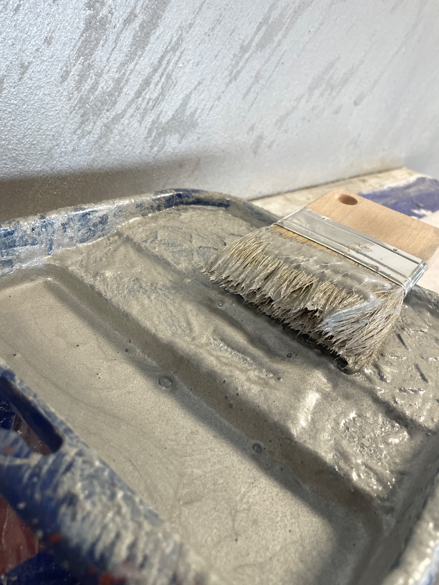 Pędzel zanurzony w szarej farbie w niebieskim kuwecie malarskim, w tle fragment ściany z efektem dekoracyjnym.