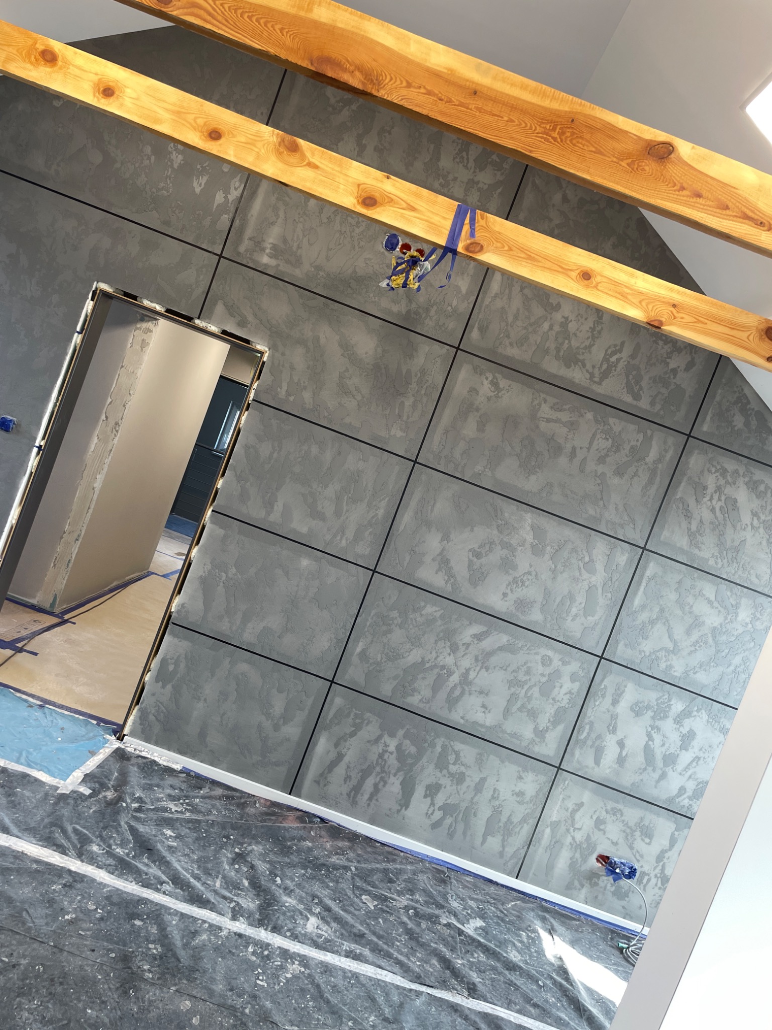 Wnętrze poddasza w trakcie prac wykończeniowych: ściana pokryta dekoracyjną szarą strukturą podzieloną na panele, widoczne drewniane belki stropowe i zabezpieczona folią podłoga.