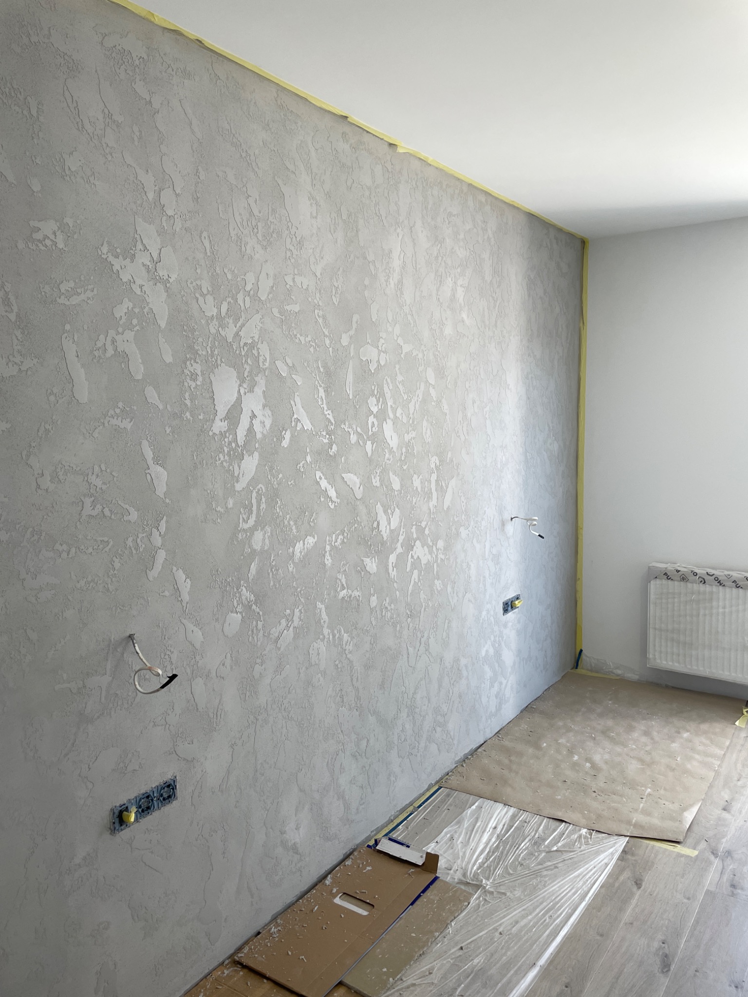 Ściana po nałożeniu dekoracyjnej, szarej struktury, zabezpieczona taśmą malarską przy suficie, z widocznymi puszkami elektrycznymi i częściowo zabezpieczoną podłogą.