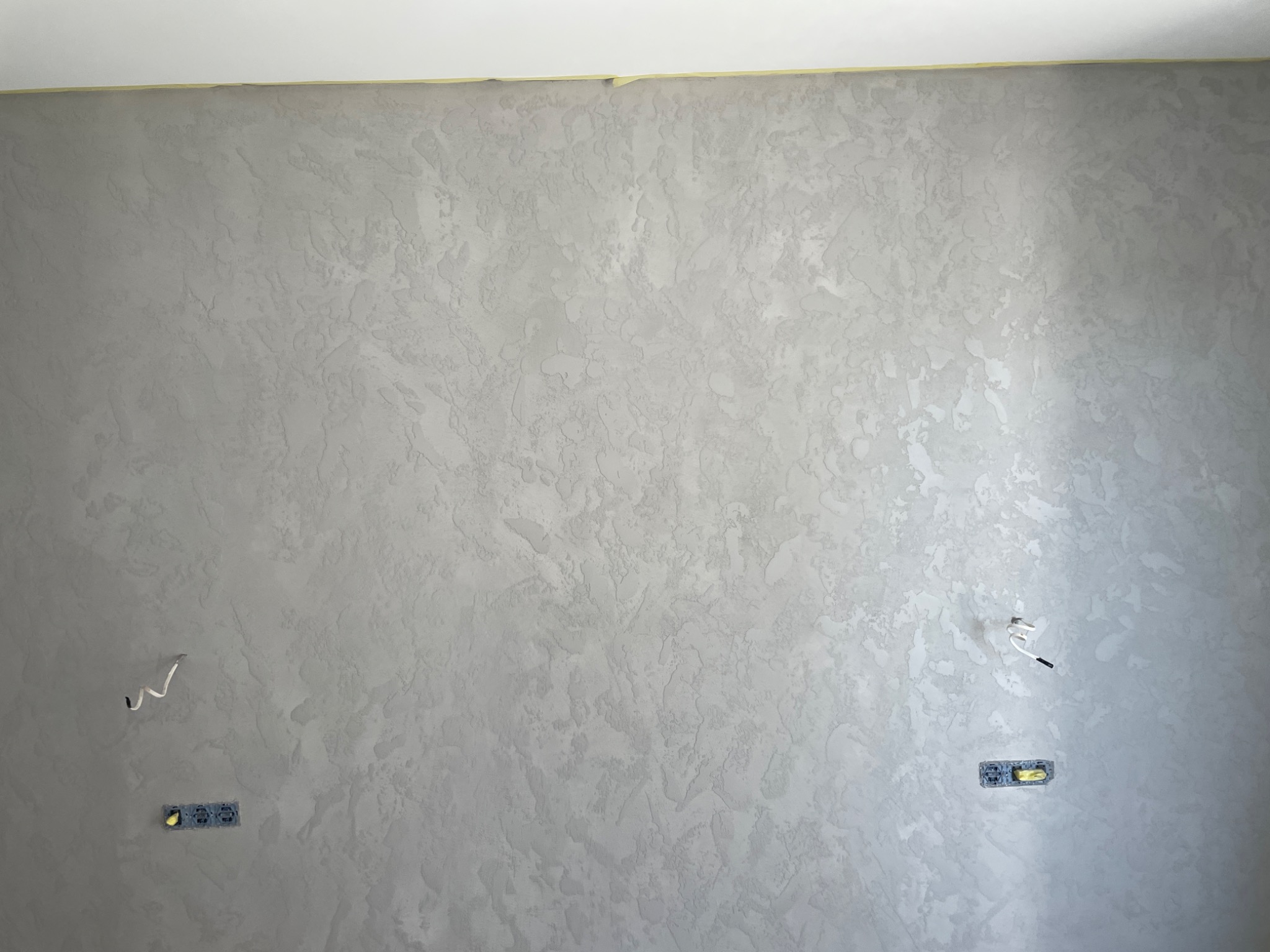 Ściana wewnętrzna z dekoracyjną szarą fakturą, zabezpieczona taśmą malarską przy suficie, widoczne gniazdka elektryczne i wystające przewody.
