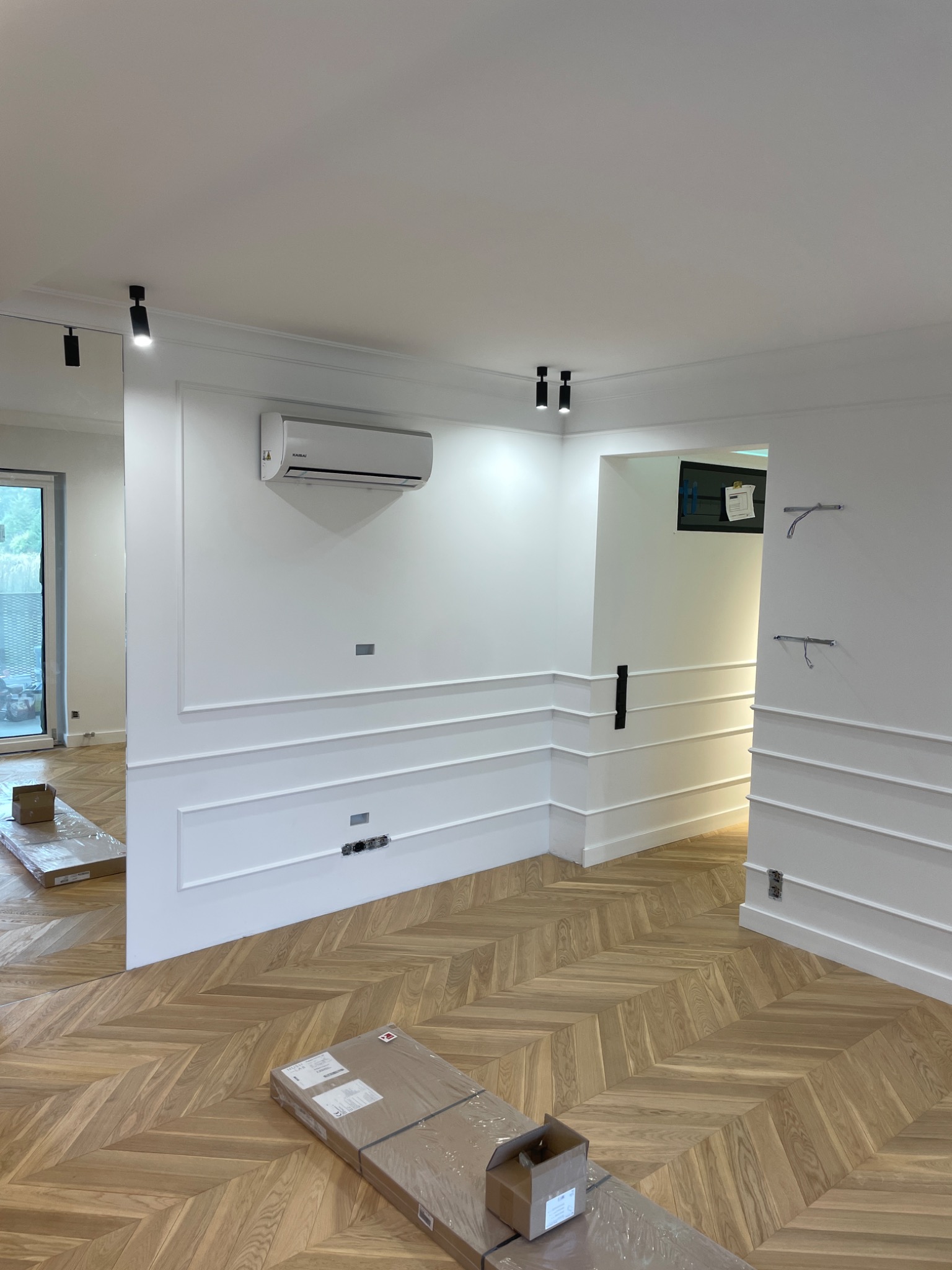 Jasne wnętrze z parkietem ułożonym we wzór jodełki francuskiej, białe ściany z ozdobnymi listwami, klimatyzator na ścianie i czarne reflektory sufitowe, paczki z panelami na podłodze.