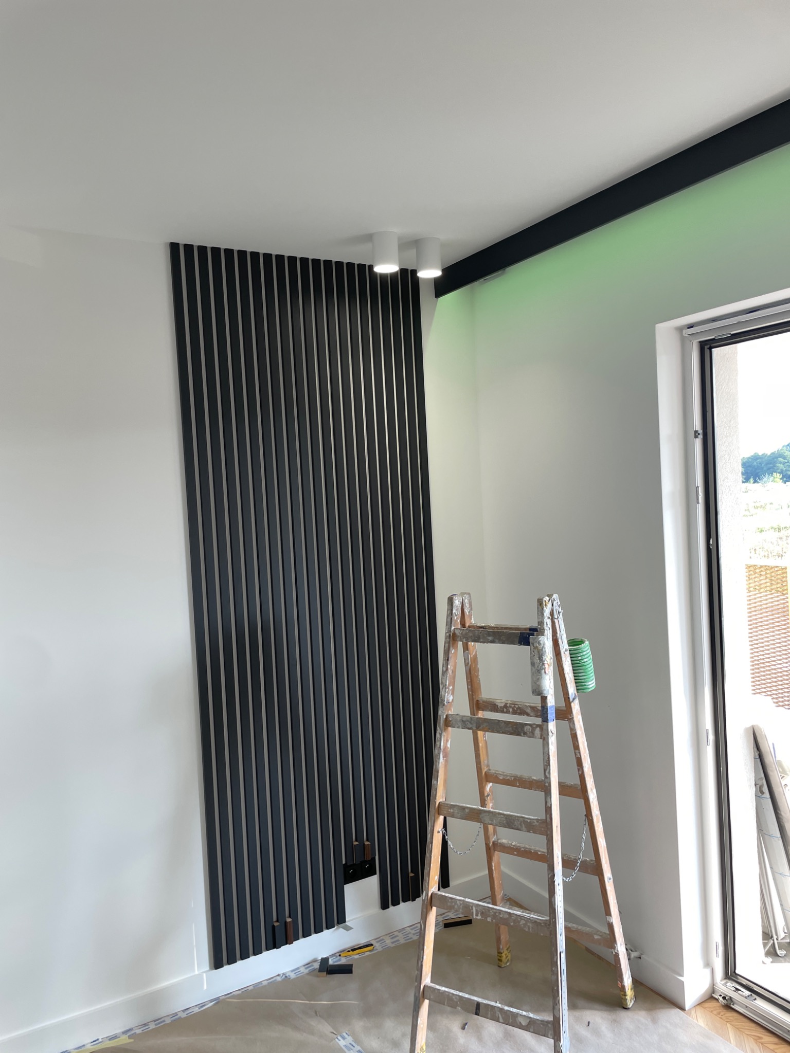 Montaż paneli dekoracyjnych ściennych w pionowe czarne listwy, widoczna drabina malarska i zabezpieczona folią podłoga.