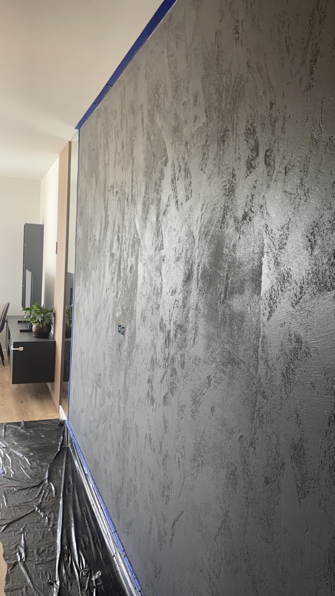 Ściana pokryta dekoracyjną szarą strukturą, zabezpieczona niebieską taśmą malarską i folią ochronną na podłodze, widoczne gniazdka elektryczne i fragment umeblowanego wnętrza.