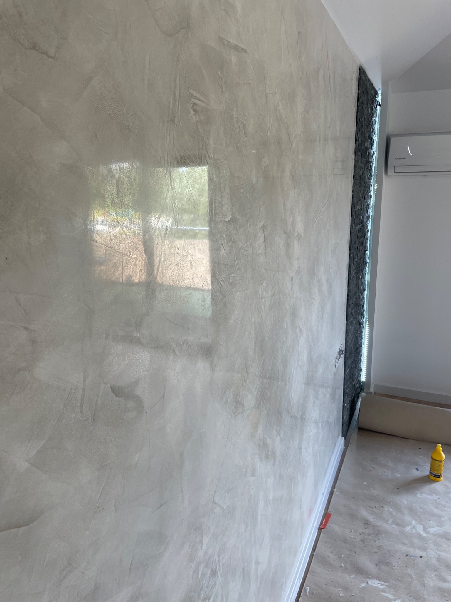 Ściana pokryta dekoracyjną, jasnoszarą strukturą z widocznym odbiciem okna, sąsiadująca z fragmentem ściany wykończonej czarnym kamieniem dekoracyjnym i klimatyzatorem; podłoga zabezpieczona folią...