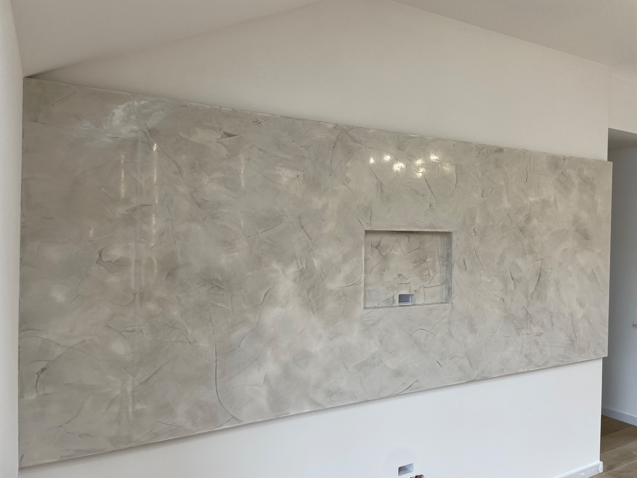 Ściana wewnętrzna wykończona dekoracyjną, szarą powłoką imitującą marmur z wnęką na element dekoracyjny, oświetlona górnym światłem.