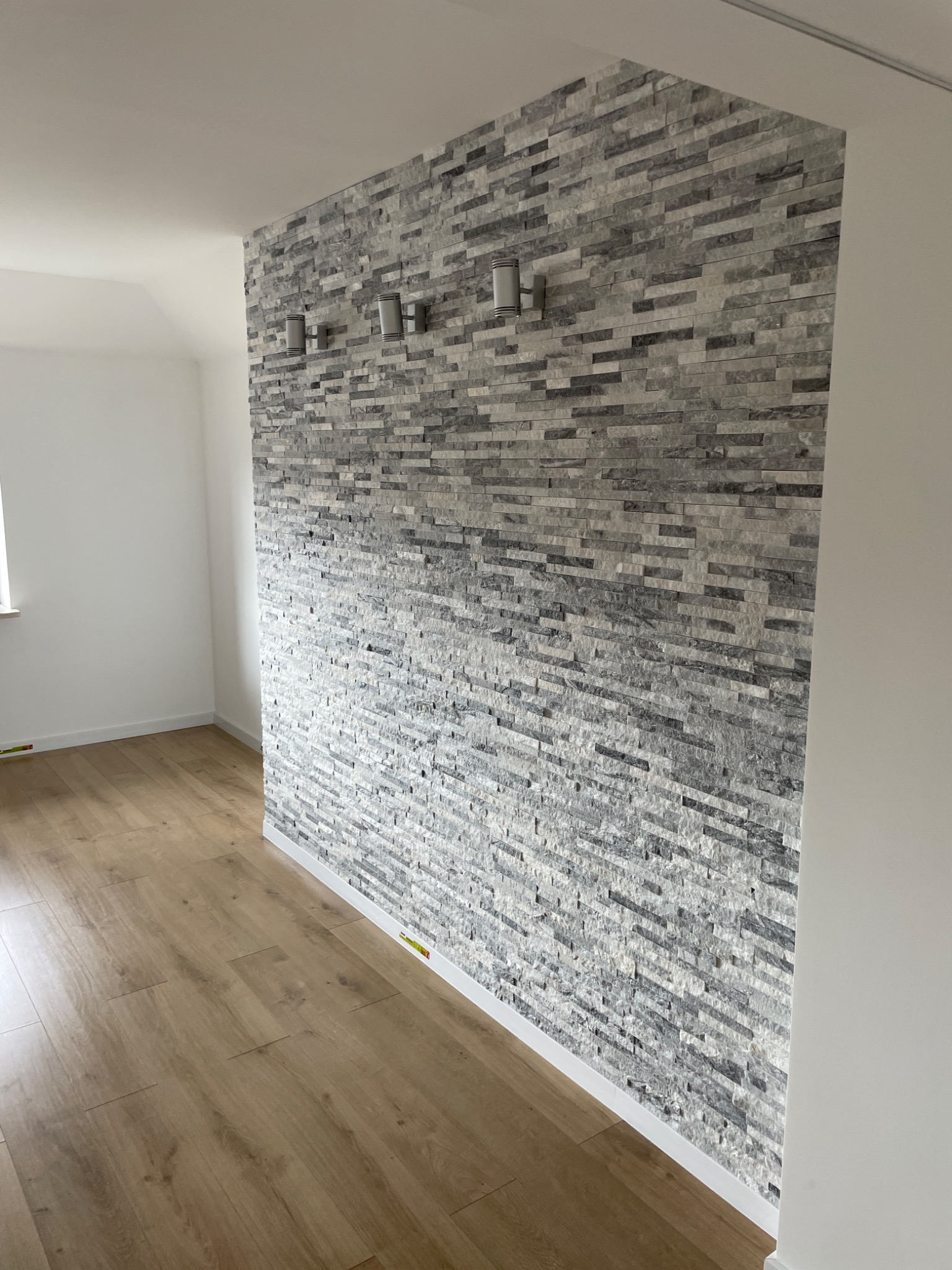 Ściana wykończona dekoracyjną okładziną z szarego kamienia, oświetlona czterema nowoczesnymi kinkietami, z widoczną podłogą z paneli o ciepłym odcieniu drewna.