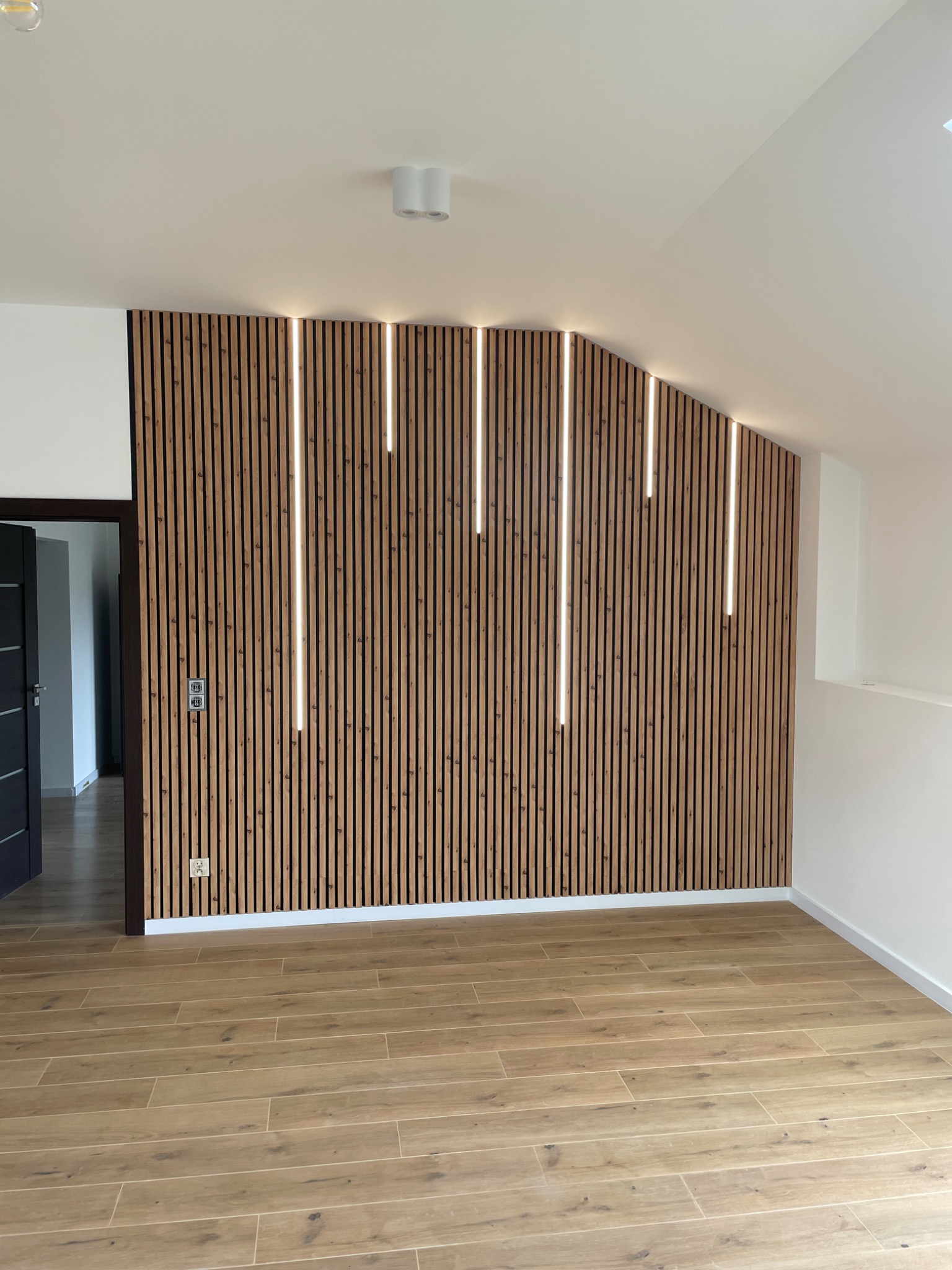 Drewniana ściana z pionowymi listwami i wbudowanym oświetleniem LED, widoczne gniazdka elektryczne, podłoga wyłożona panelami imitującymi drewno, drzwi wejściowe po lewej stronie.