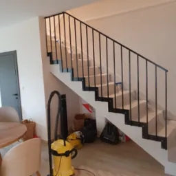 Nowoczesne schody z czarną, metalową balustradą w jasnym wnętrzu, zabezpieczone folią ochronną na czas remontu.