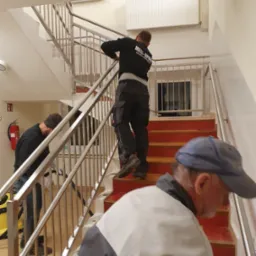 Montaż balustrady ze stali nierdzewnej na schodach w budynku, widoczni trzej pracownicy podczas pracy, jeden z nich w bluzie z logo firmy.