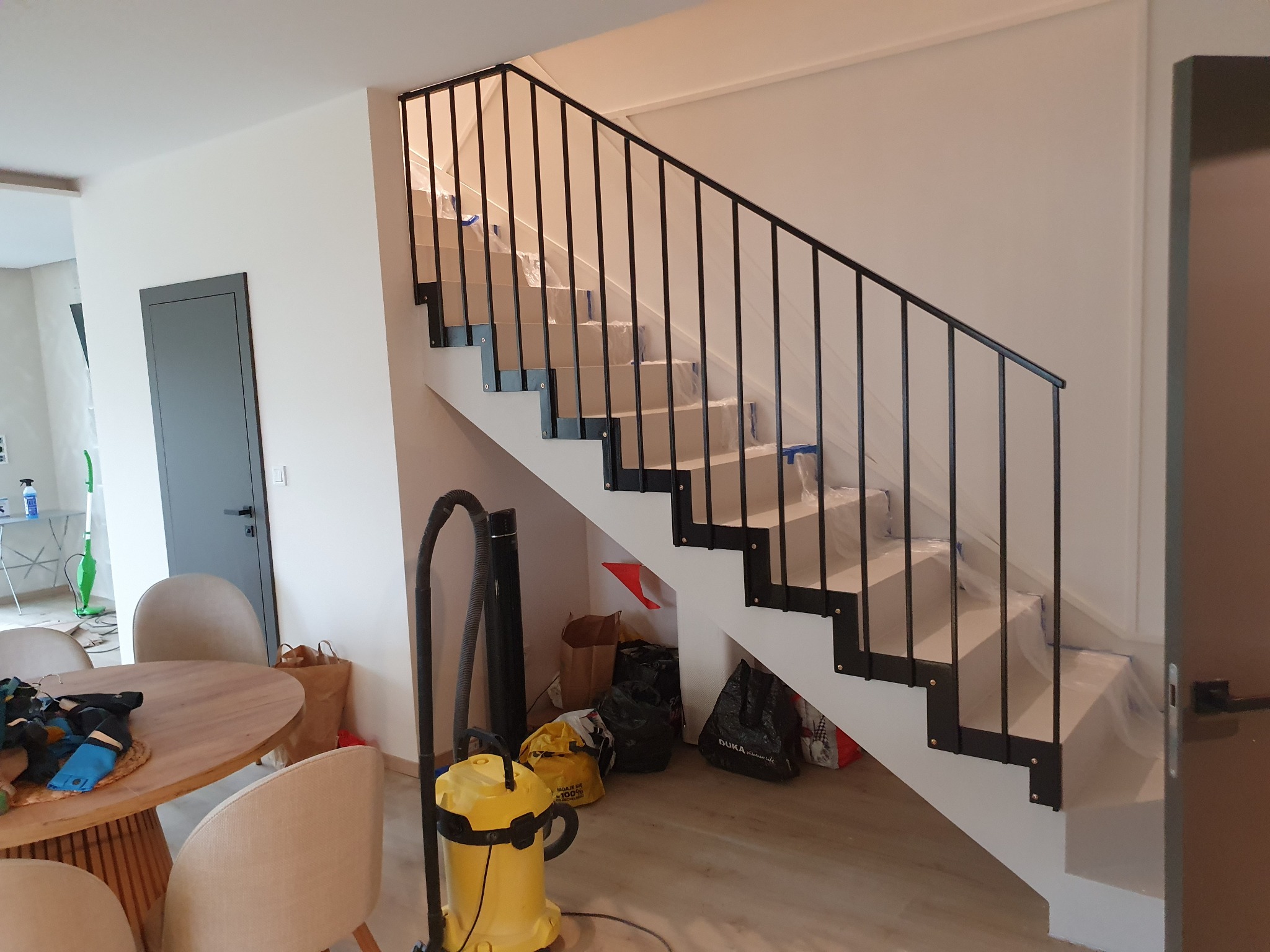 Nowoczesne schody z czarną, metalową balustradą w jasnym wnętrzu, zabezpieczone folią ochronną na czas remontu.