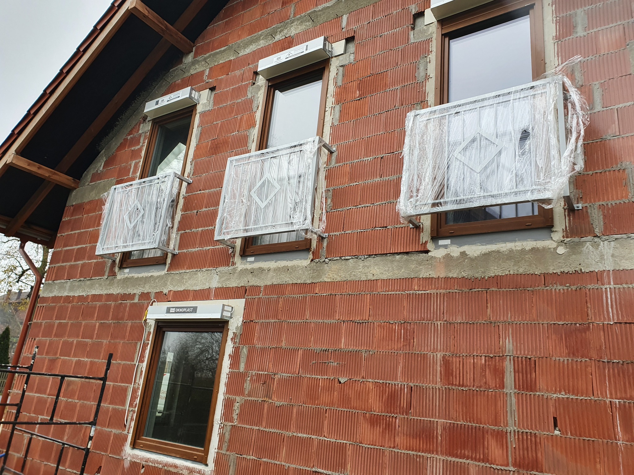 Nowe, jeszcze ofoliowane balustrady okienne z metalowymi wzorami, zamontowane na surowej elewacji budynku z czerwonej cegły kratówki.