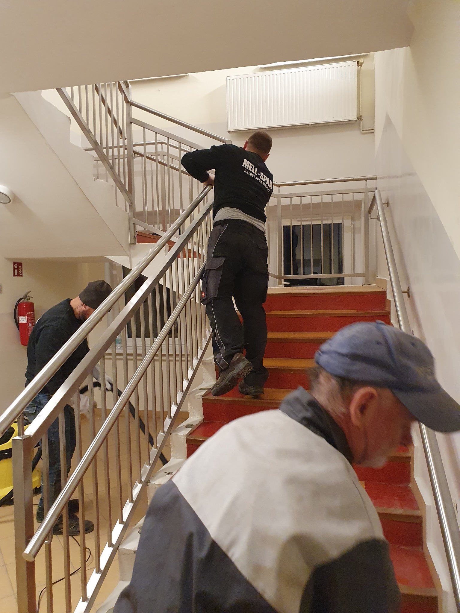 Montaż balustrady ze stali nierdzewnej na schodach w budynku, widoczni trzej pracownicy podczas pracy, jeden z nich w bluzie z logo firmy.