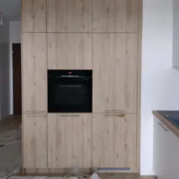 Wysoka zabudowa kuchenna z frontami w kolorze jasnego drewna i wbudowanym piekarnikiem, zabezpieczona folią malarską na podłodze.