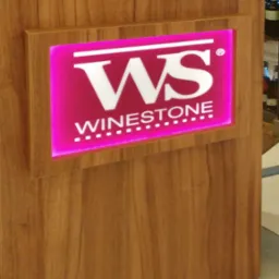 Drewniana lada recepcyjna z podświetlanym logo Winestone w kolorze różowym, widoczne butelki w tle.