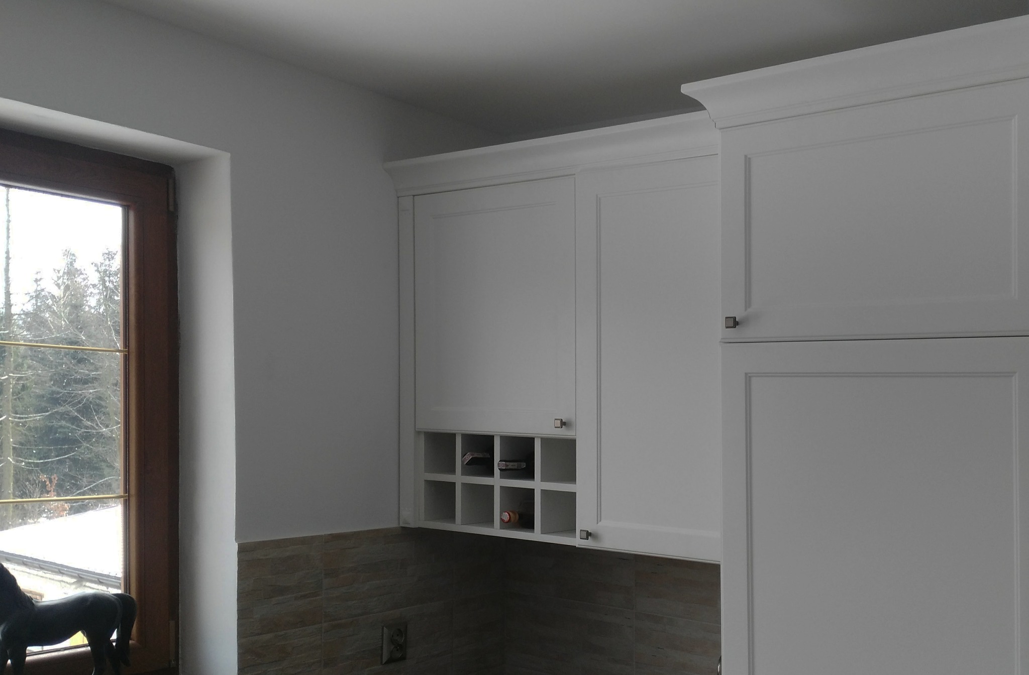 Białe szafki kuchenne z górnymi szafkami, w tym szafka z przegródkami na butelki, widok z bliska na tle okna z widokiem na drzewa.