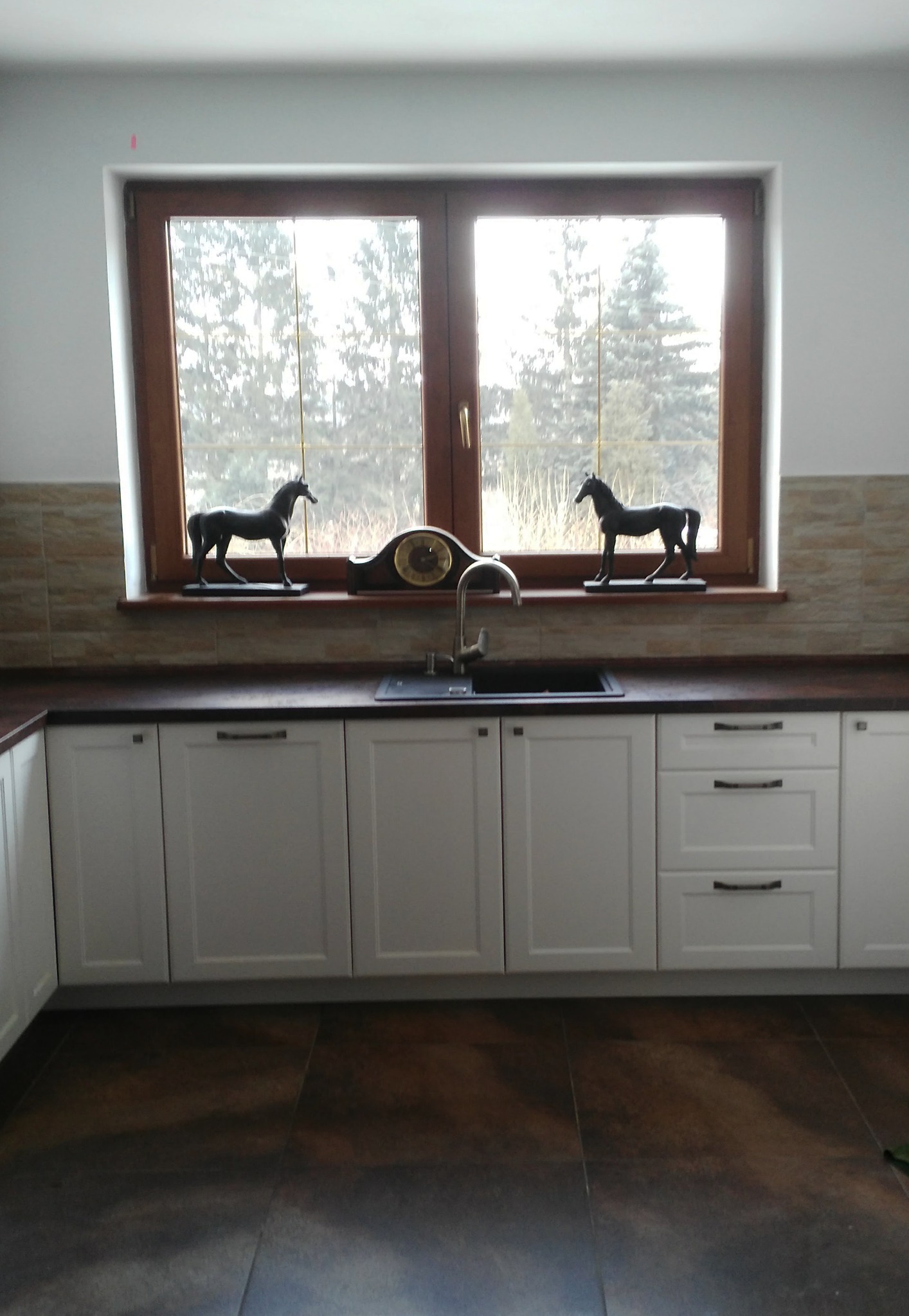 Białe szafki kuchenne z ciemnym blatem i dekoracjami okiennymi: figurki koni i zegar.