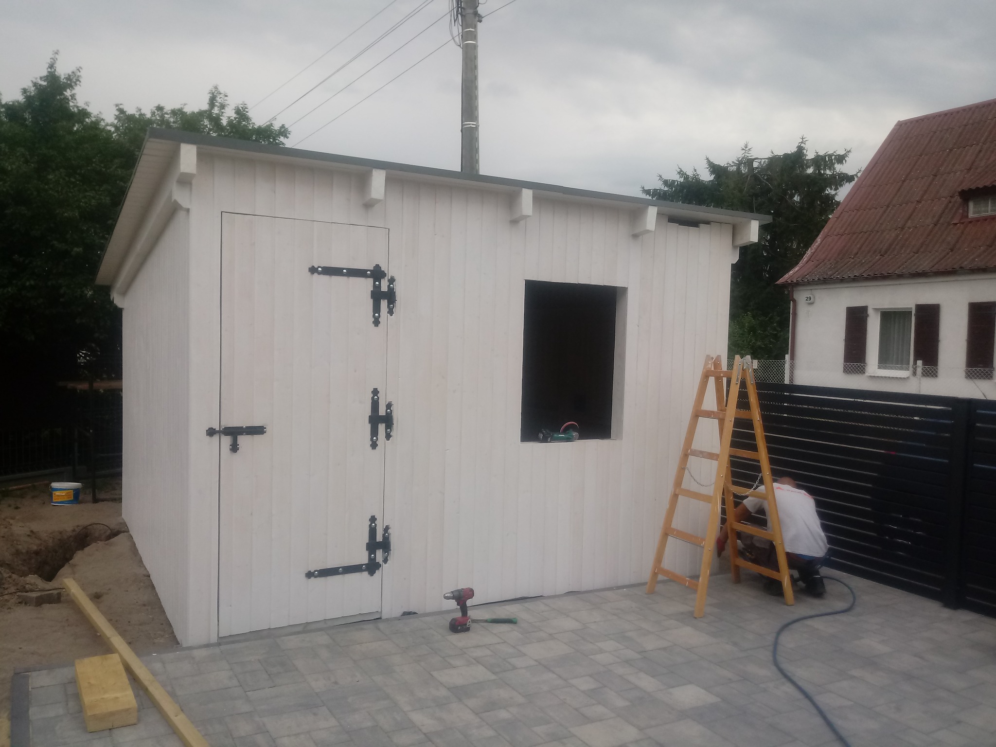 Biały, drewniany budynek gospodarczy z czarnym, kutym okuciem drzwiowym, w trakcie budowy okna, z widocznymi narzędziami i pracownikiem malującym ogrodzenie w tle.