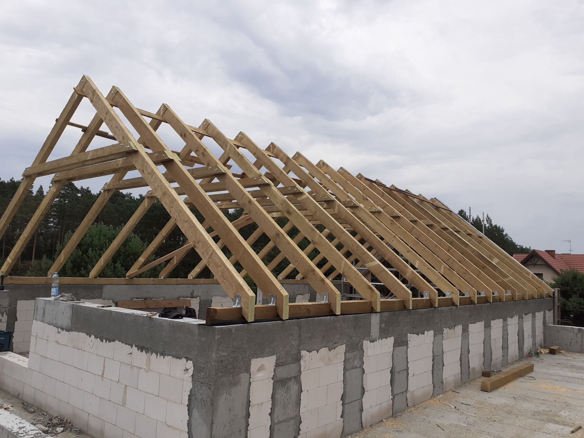 Surowa konstrukcja drewniana dachu z widocznymi wiązaniami i elementami mocującymi, wznoszona na murowanych ścianach budynku w stanie surowym otwartym, na tle pochmurnego nieba i zieleni drzew.