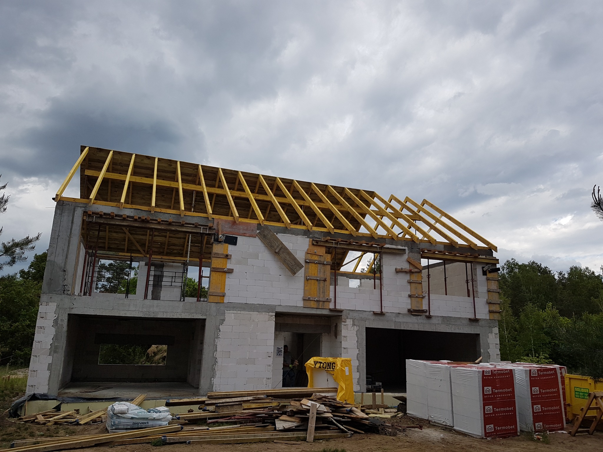 Budowa domu jednorodzinnego w stanie surowym otwartym, widoczna więźba dachowa z żółtych belek, ściany z białych bloczków, na ziemi materiały budowlane i żółty worek YTONG.