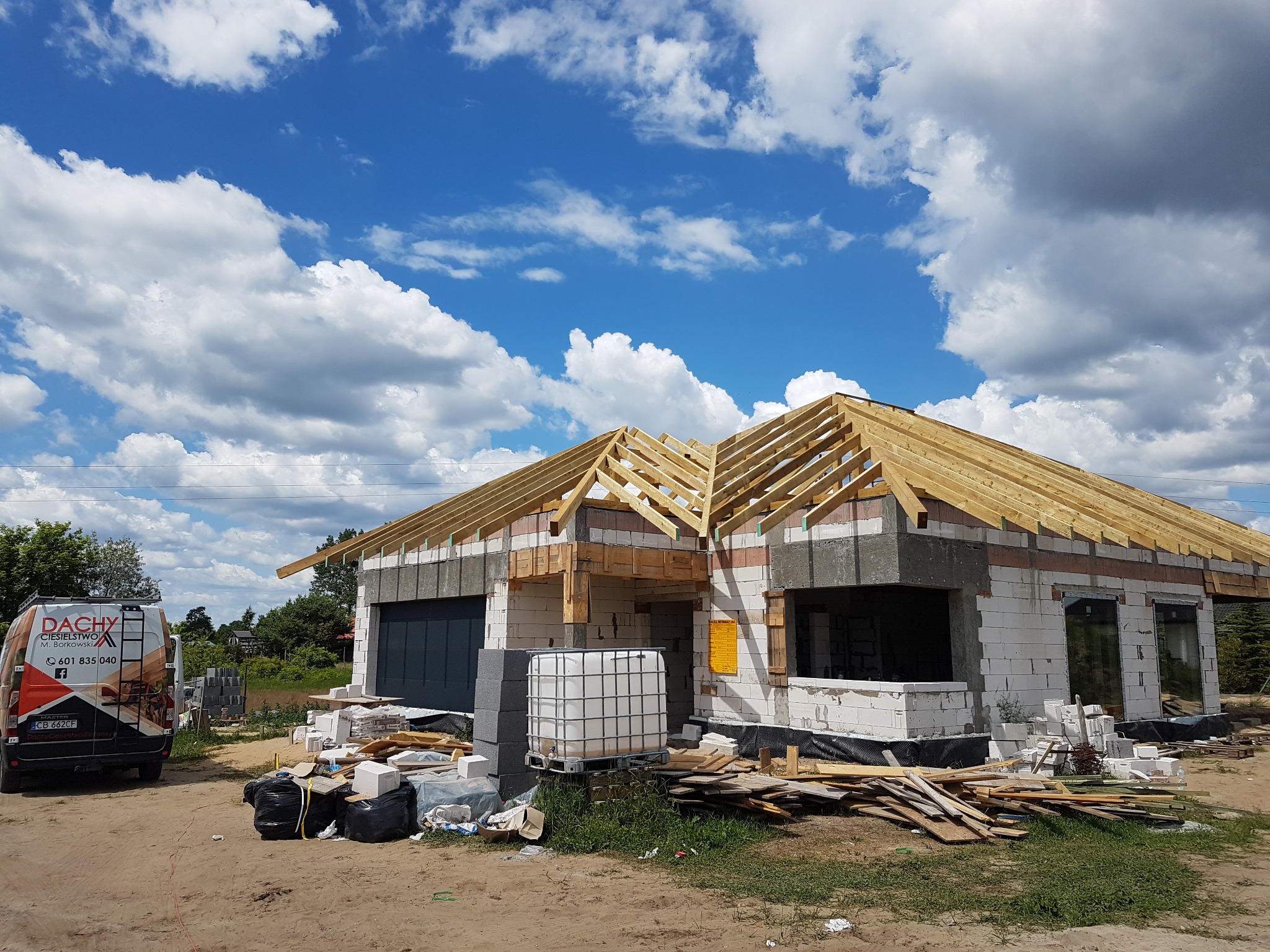 Budowa domu jednorodzinnego w trakcie montażu drewnianej konstrukcji dachu, widoczny samochód dostawczy z logo firmy dekarskiej na tle błękitnego nieba z chmurami.