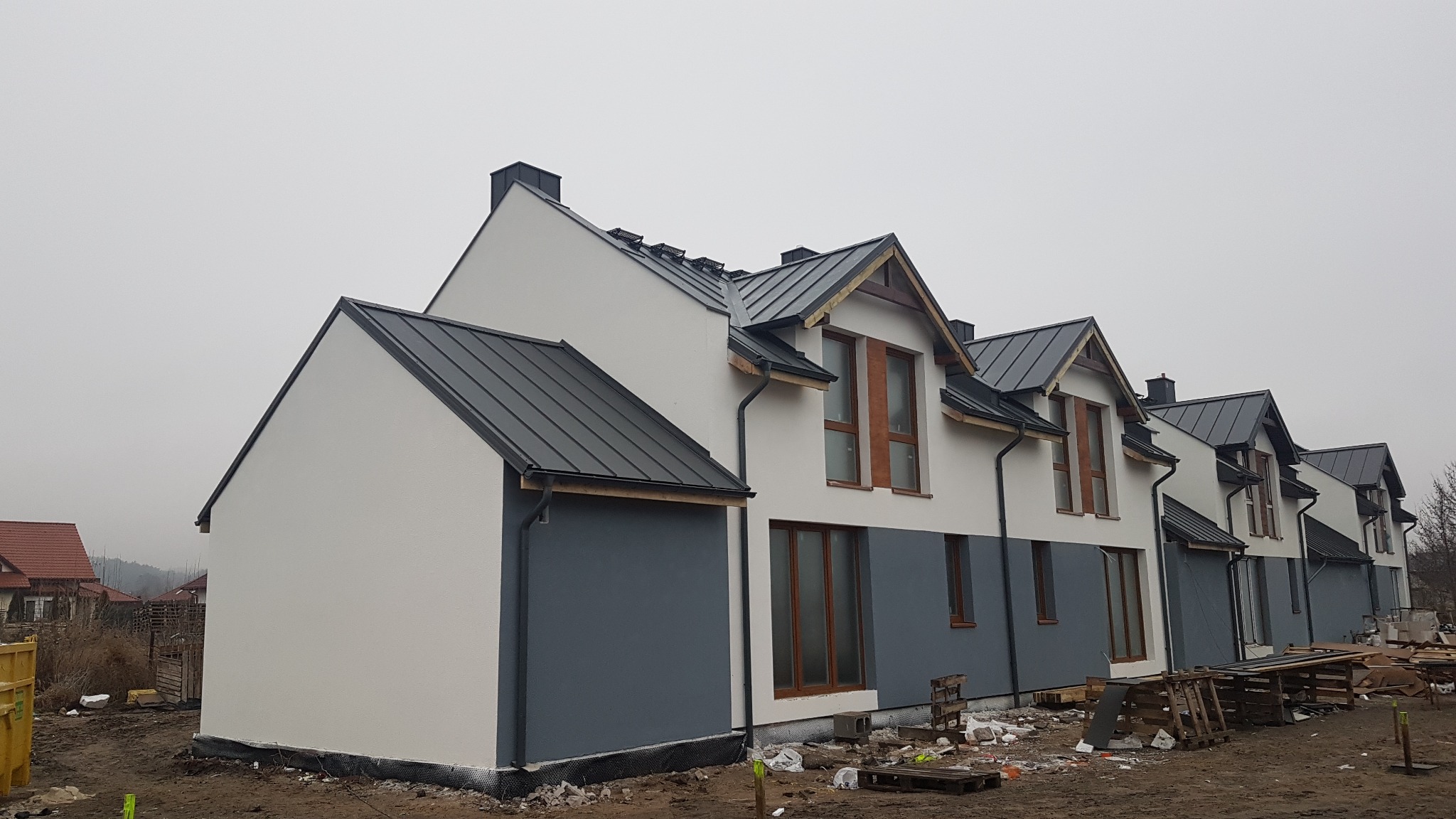 Szereg nowych domów jednorodzinnych z dwuspadowymi dachami krytymi blachą na rąbek stojący, widoczne rynny i obróbki blacharskie, elewacja w kolorach bieli i szarości, teren wokół w trakcie...