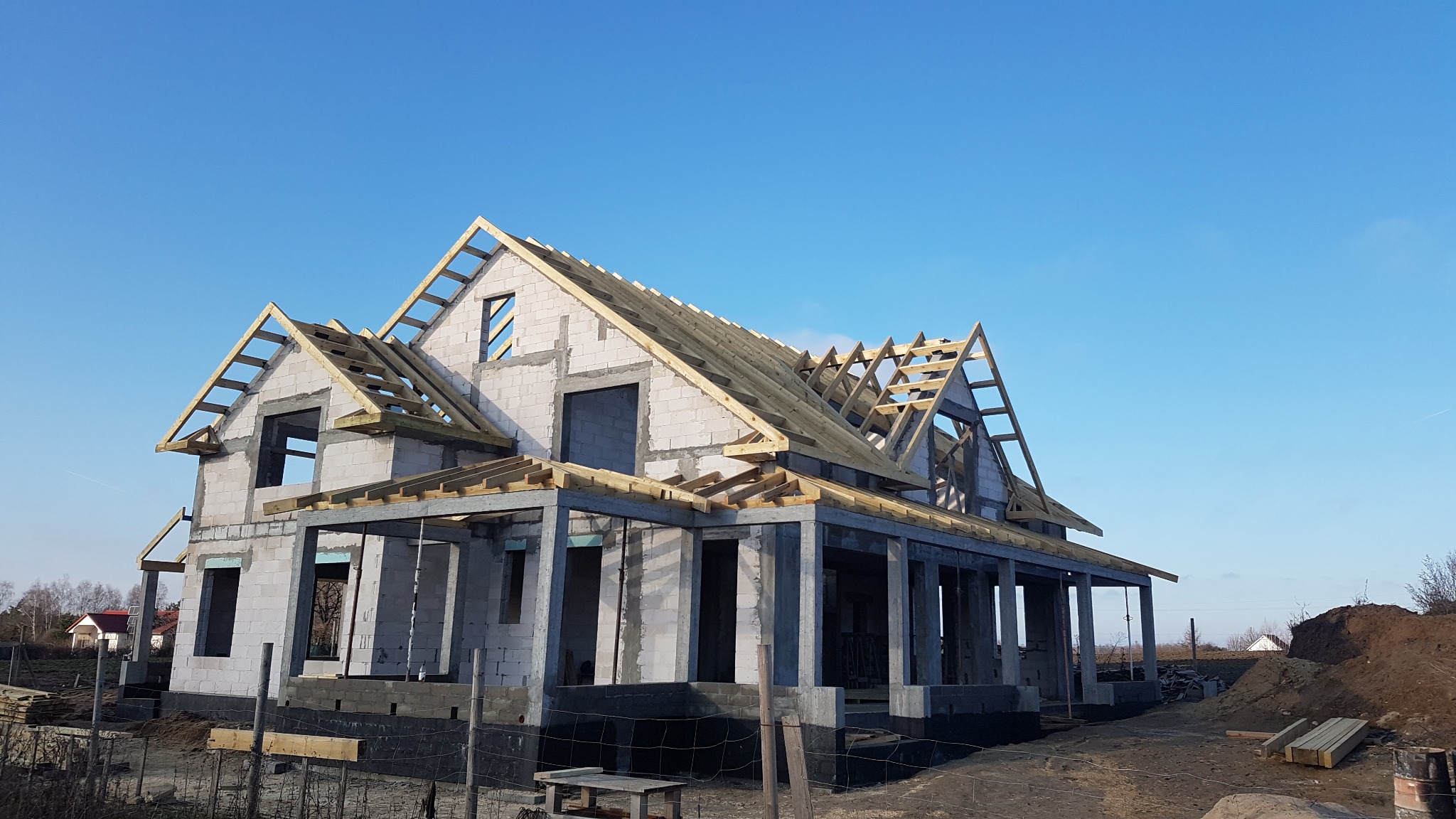 Surowy stan domu jednorodzinnego z białych bloczków, w trakcie budowy drewnianej konstrukcji dachu na tle błękitnego nieba.