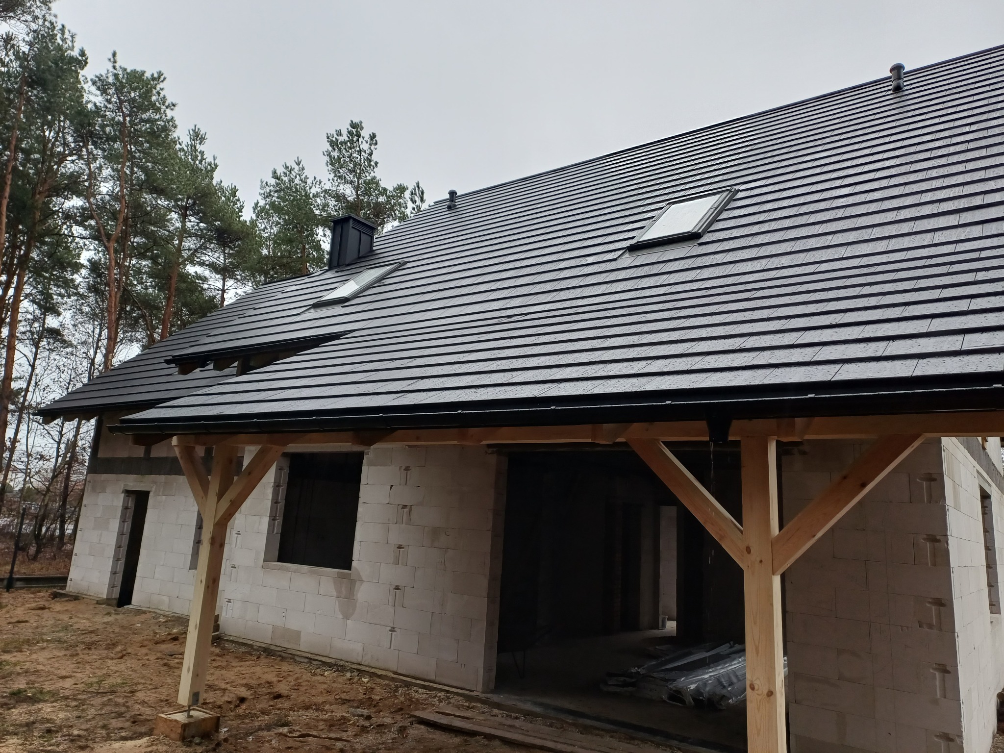 Nowoczesny dom w trakcie budowy z ciemną dachówką, widoczne okna dachowe, komin i drewniana konstrukcja werandy na tle drzew i pochmurnego nieba.