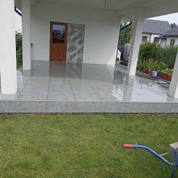 Usługi Bruk-Bud JUKO YURII KOVALOV - Wykończony taras z szarych płytek ceramicznych, widok z poziomu trawnika, z fragmentem taczki z ziemią w rogu kadru.