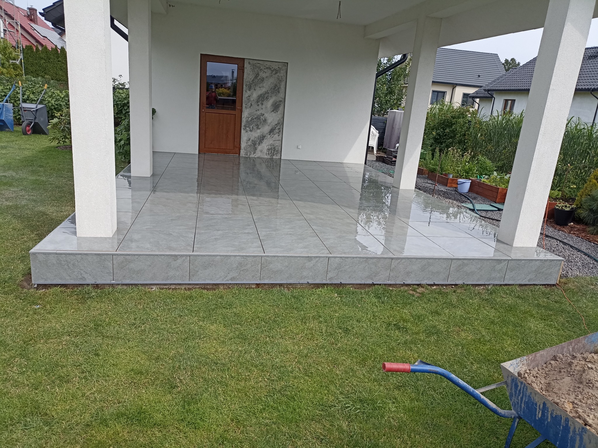 Wykończony taras z szarych płytek ceramicznych, widok z poziomu trawnika, z fragmentem taczki z ziemią w rogu kadru.