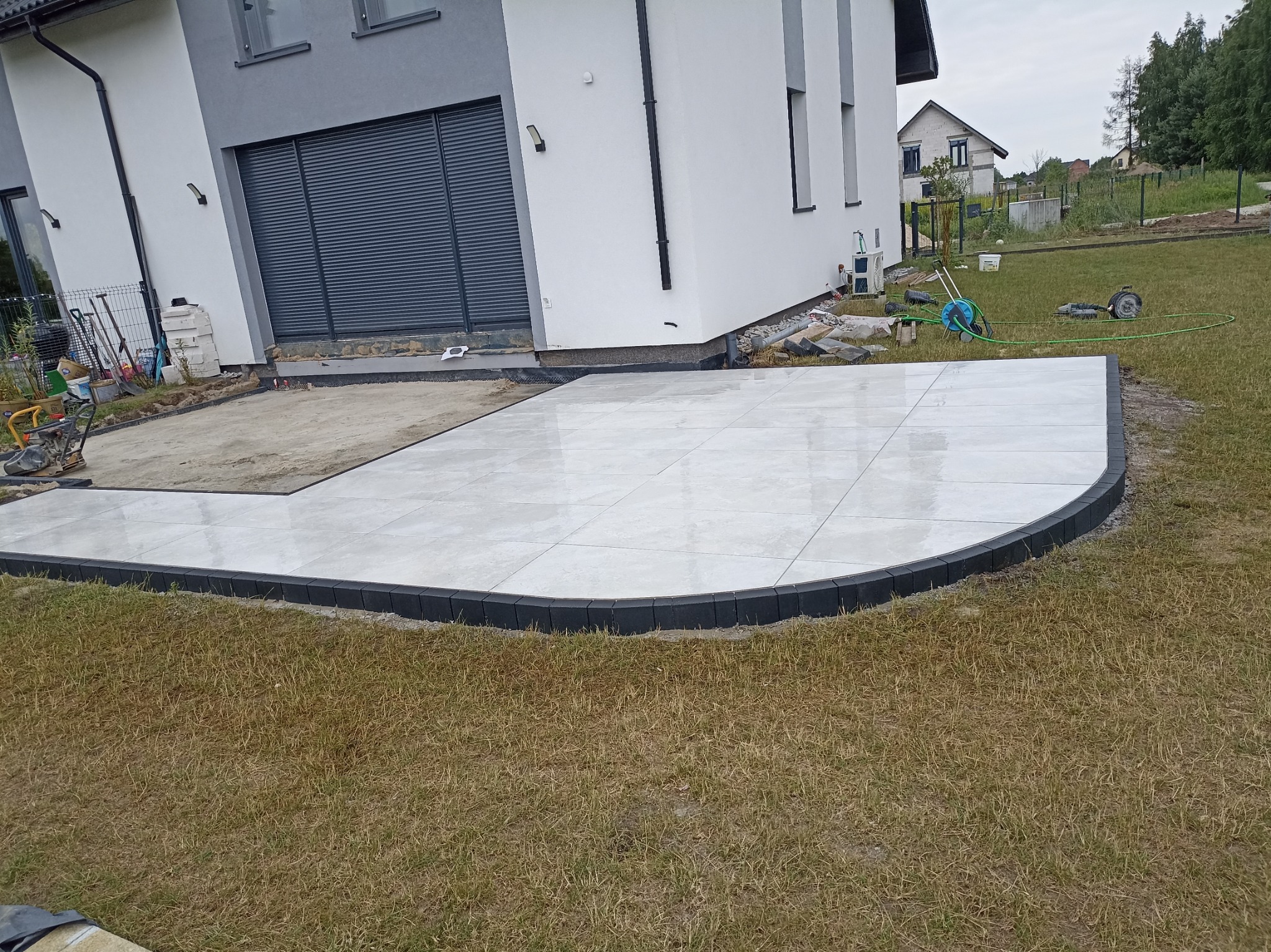 Wyłożony jasnoszarymi płytami taras przylegający do nowoczesnego domu z szarą stolarką okienną, obrzeża tarasu wykończone ciemnymi kostkami, widoczne narzędzia i materiały budowlane w tle.