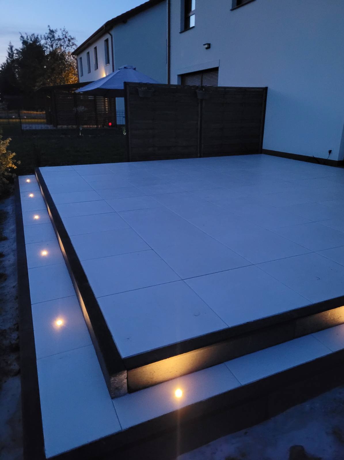 Nowoczesny taras wyłożony jasnymi płytami w wieczornym oświetleniu, z niskim ogrodzeniem w tle i subtelnymi punktowymi lampami LED wzdłuż krawędzi.
