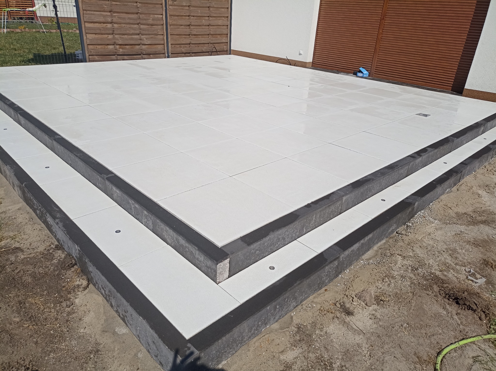 Wykończony taras z białych, kwadratowych płyt ułożonych na dwóch poziomach schodów z ciemnoszarą obwódką, widoczne punkty świetlne w podstopnicach, w tle drewniane ogrodzenie i fragment budynku.