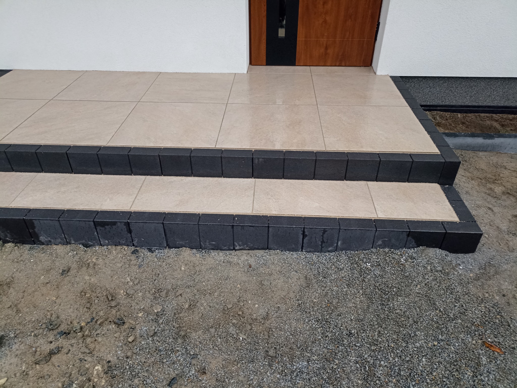 Wykończone dwustopniowe schody wejściowe do domu we Wrocławiu, wyłożone jasnymi płytkami z ciemną obwódką, widok z poziomu gruntu na świeżo obsypane kamieniami podłoże.