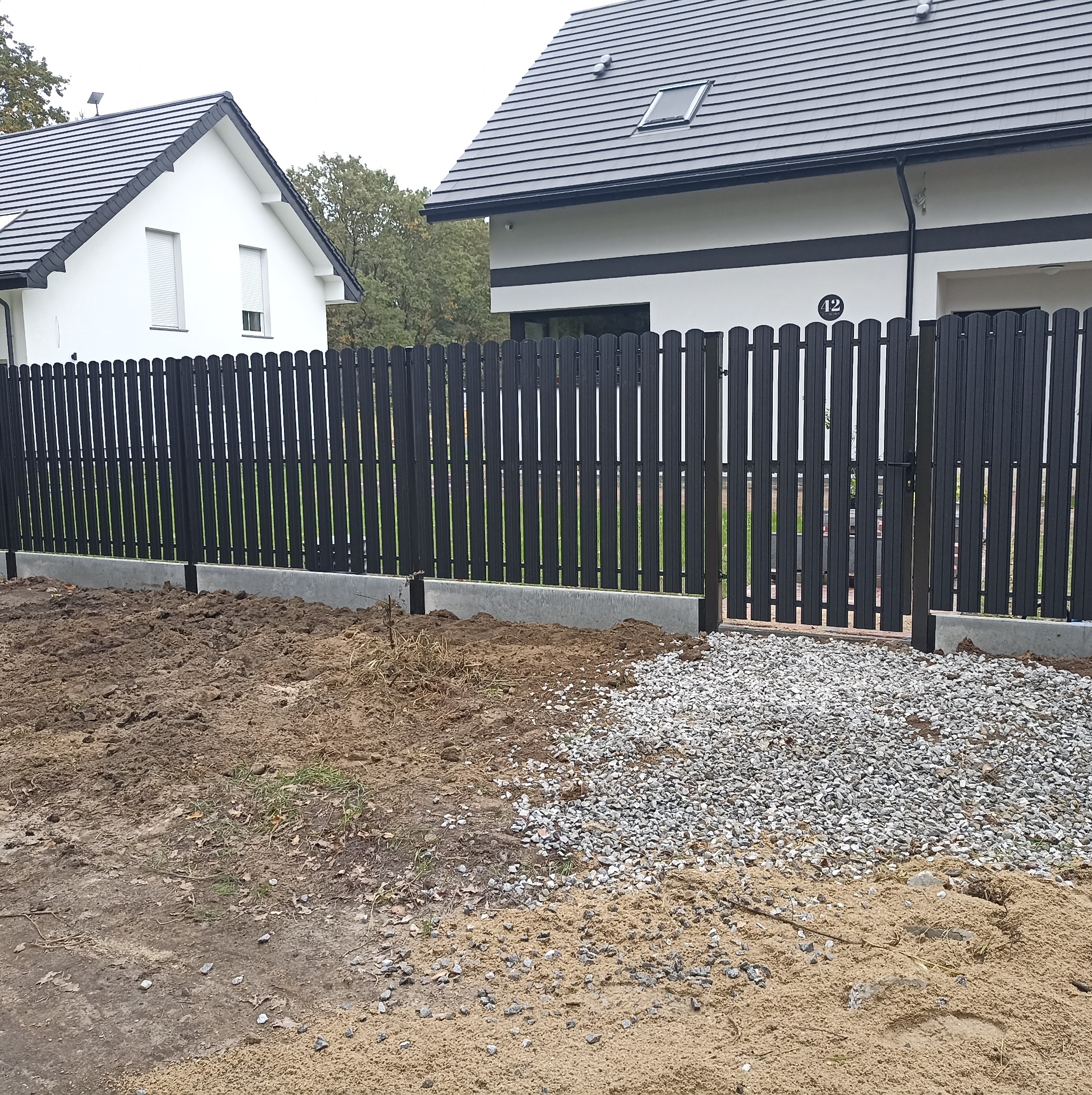 Nowoczesne, czarne ogrodzenie panelowe z furtką, na betonowej podmurówce, przed nowo wybudowanymi domami z szarą dachówką, teren wokół ogrodzenia częściowo wyrównany żwirem i piaskiem.