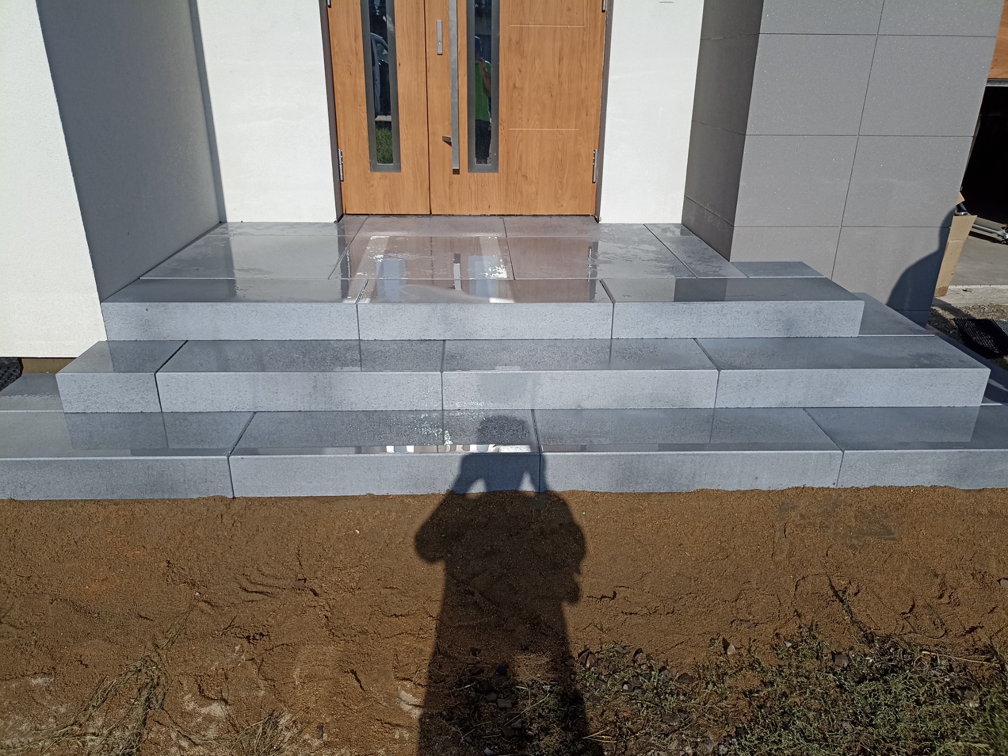 Schody wejściowe do domu wyłożone szarą kostką brukową, widoczny cień osoby fotografującej na ziemi.