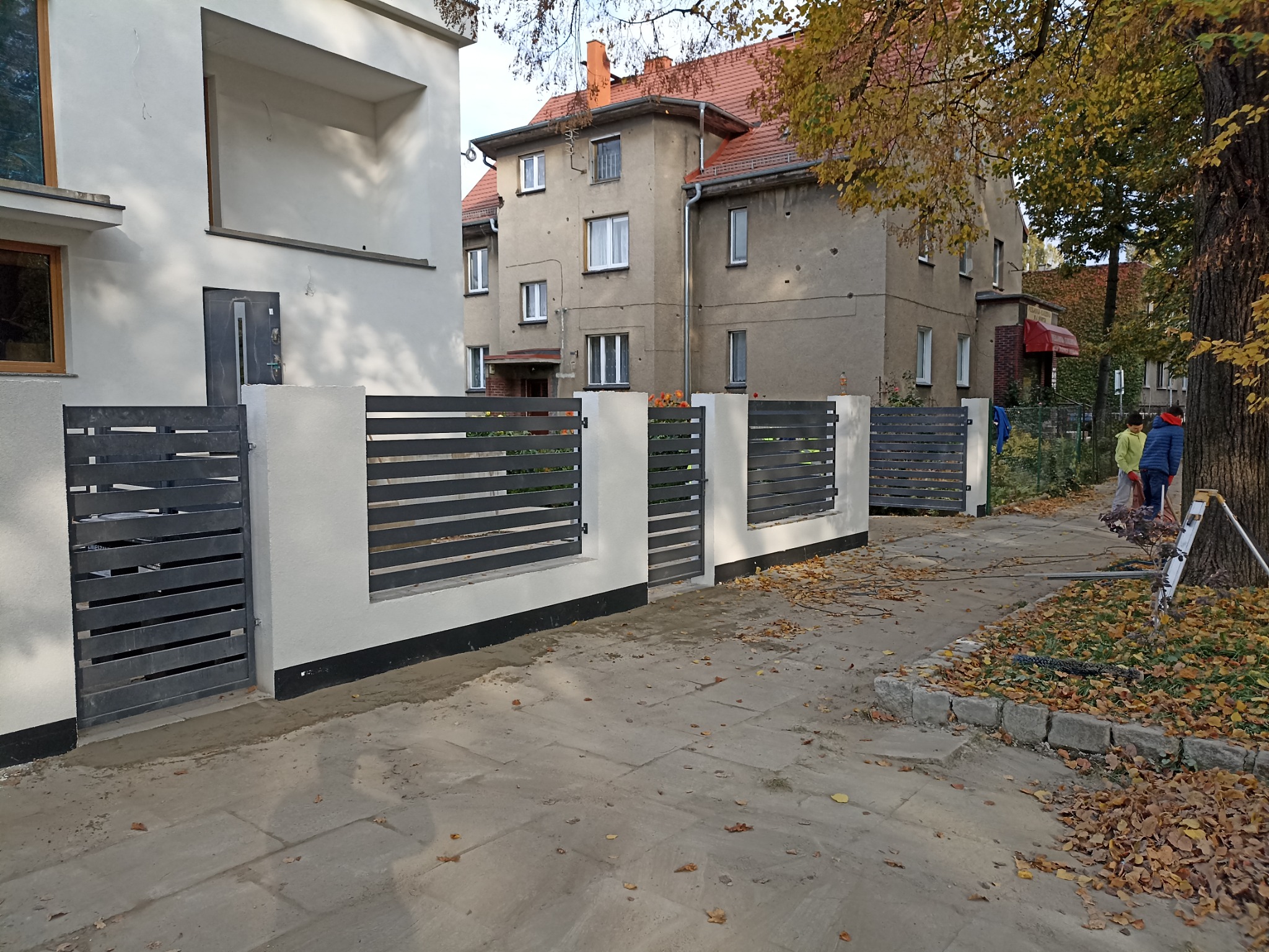 Nowoczesne ogrodzenie z poziomych, szarych paneli metalowych na białych słupkach, widok z zewnątrz posesji, fragment chodnika z opadłymi liśćmi