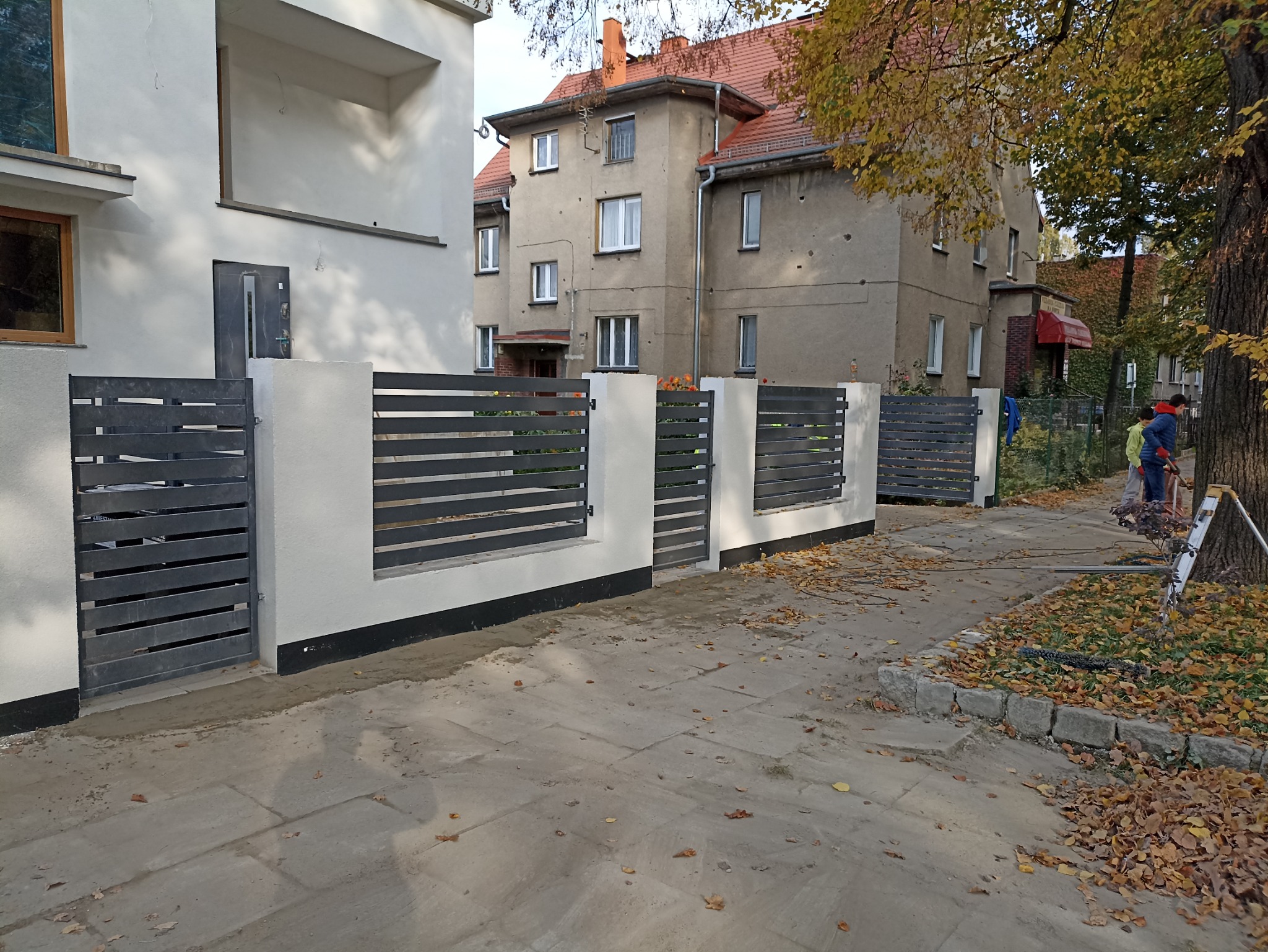 Nowoczesne ogrodzenie z poziomych, grafitowych paneli zamontowane na białych, betonowych słupkach, oddzielające posesję od chodnika w jesiennej scenerii.