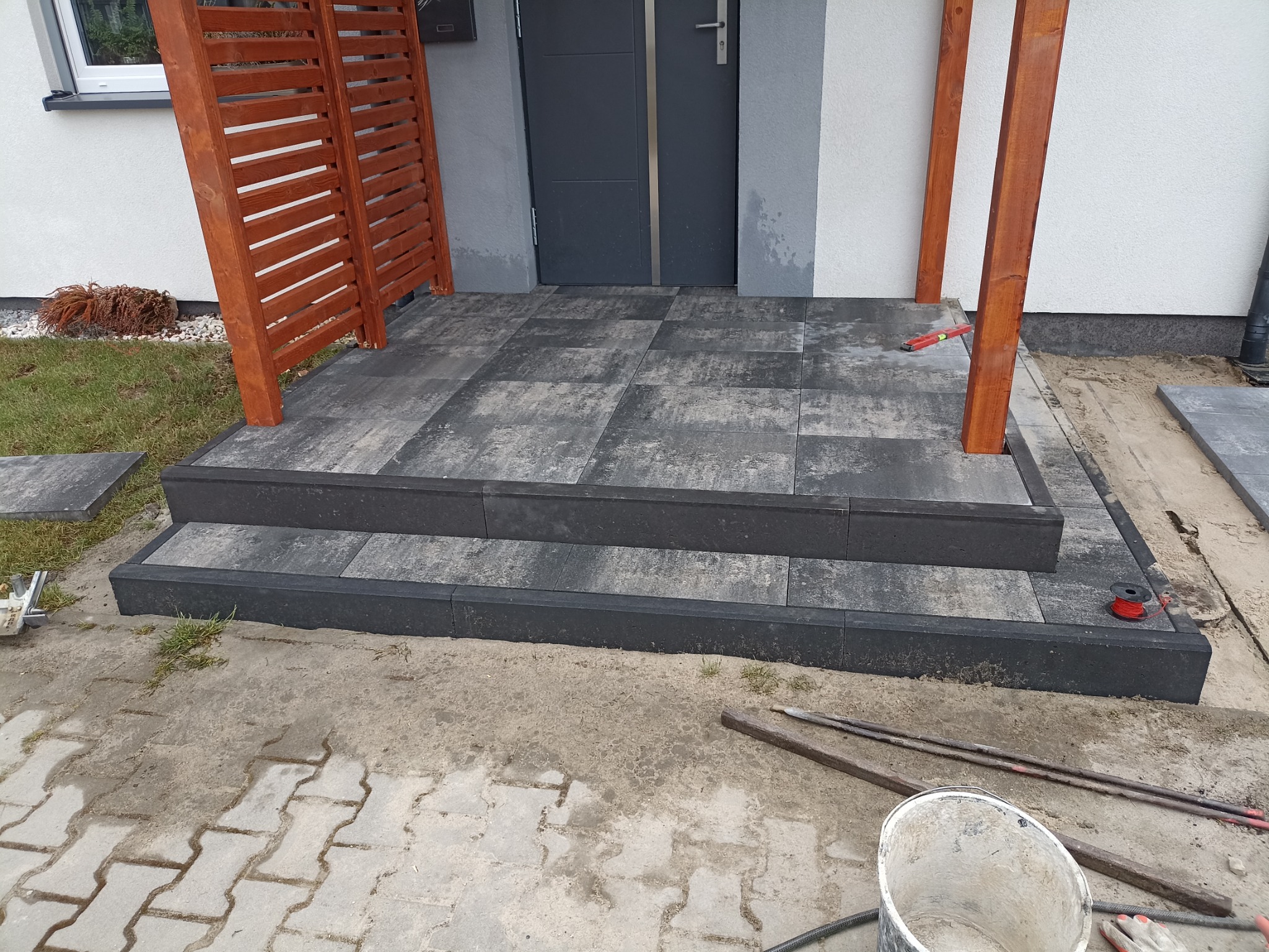 Schody wejściowe wyłożone szarą kostką brukową z drewnianą konstrukcją zadaszenia, widoczne narzędzia i materiały budowlane w trakcie prac wykończeniowych.