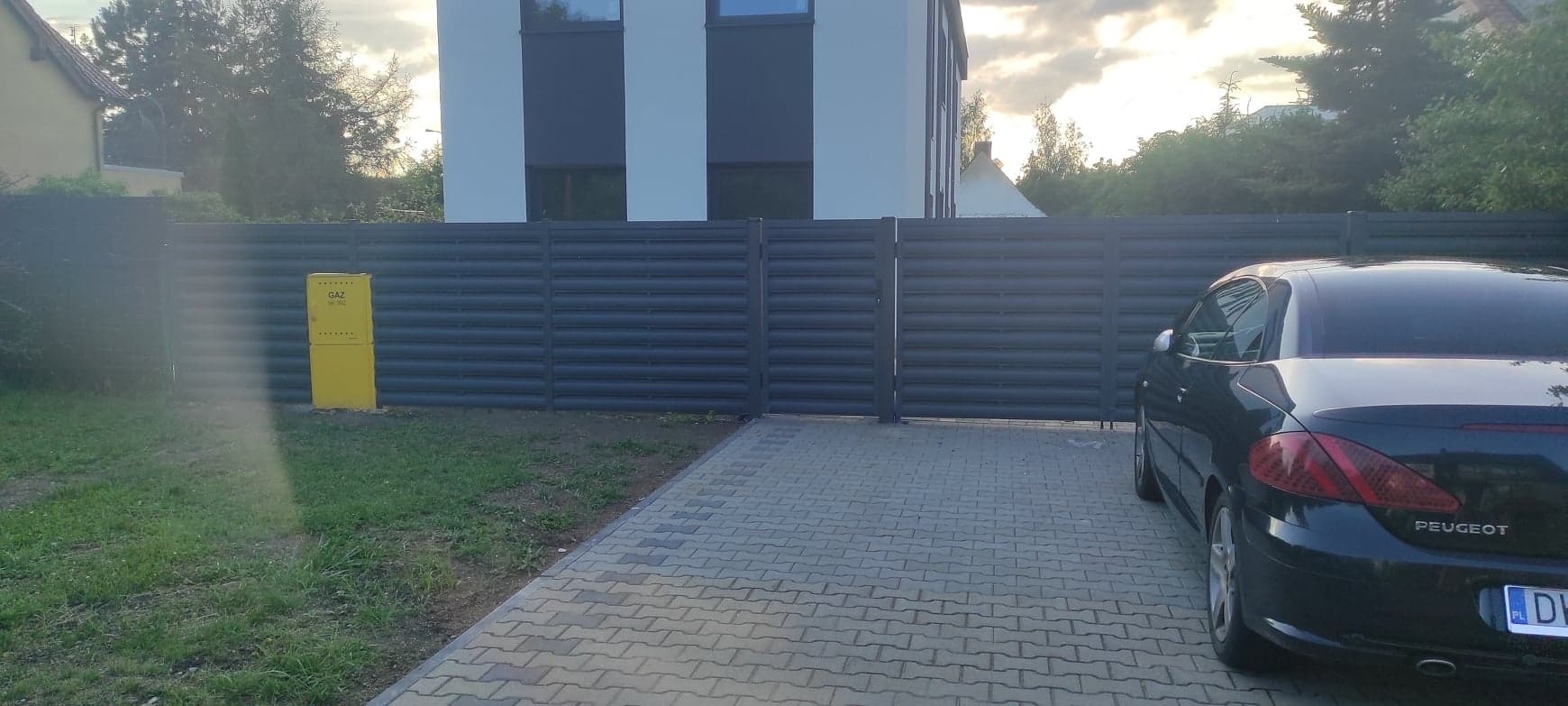 Nowoczesne ogrodzenie panelowe w kolorze grafitowym z bramą wjazdową, widoczny żółty gazomierz i zaparkowany samochód osobowy na kostce brukowej, tło stanowi budynek mieszkalny i zieleń.